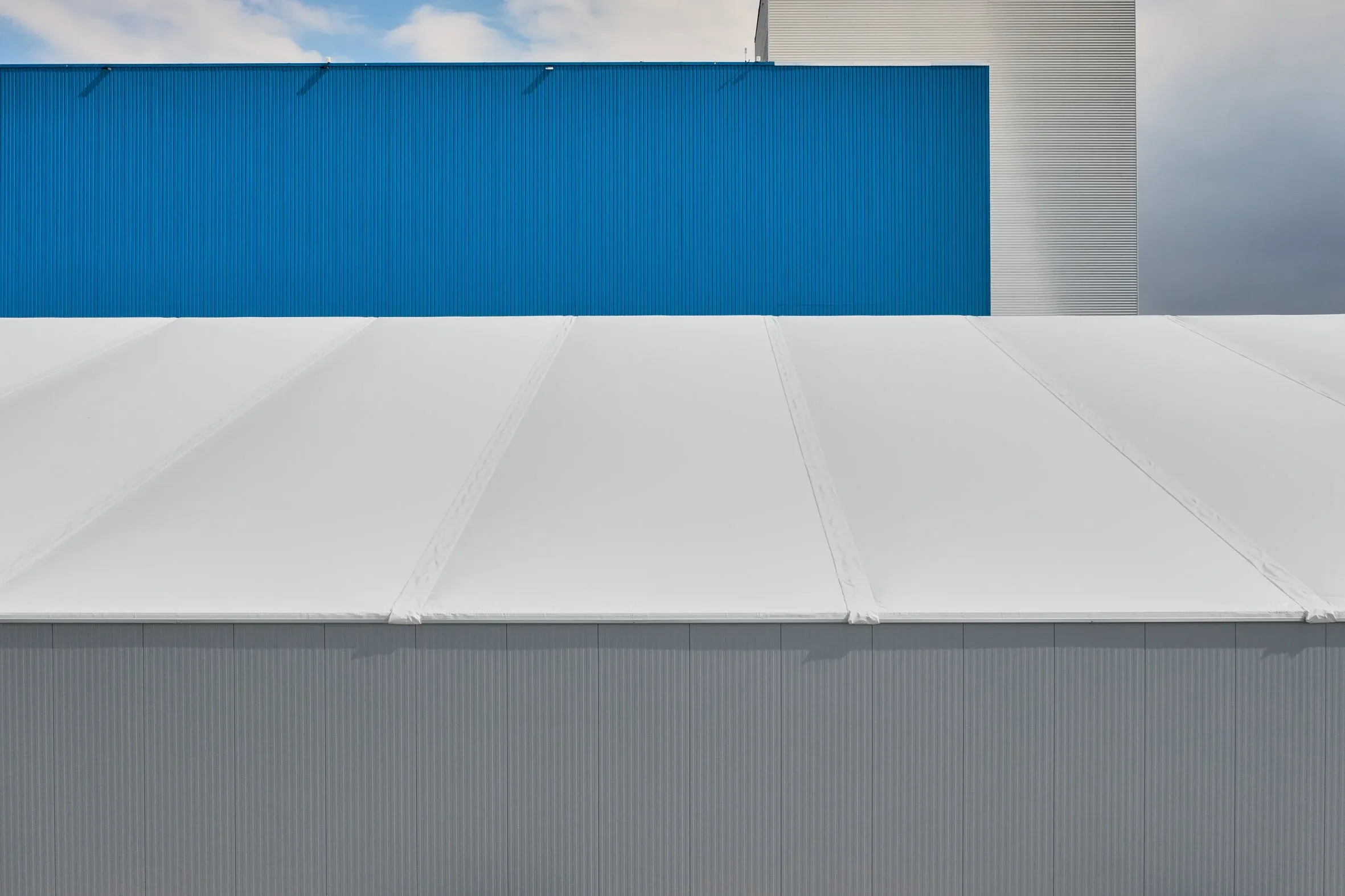 Außendetail, weiße Membranhülle und blaue Profilfassade einer Halle vor leicht bewölktem Himmel