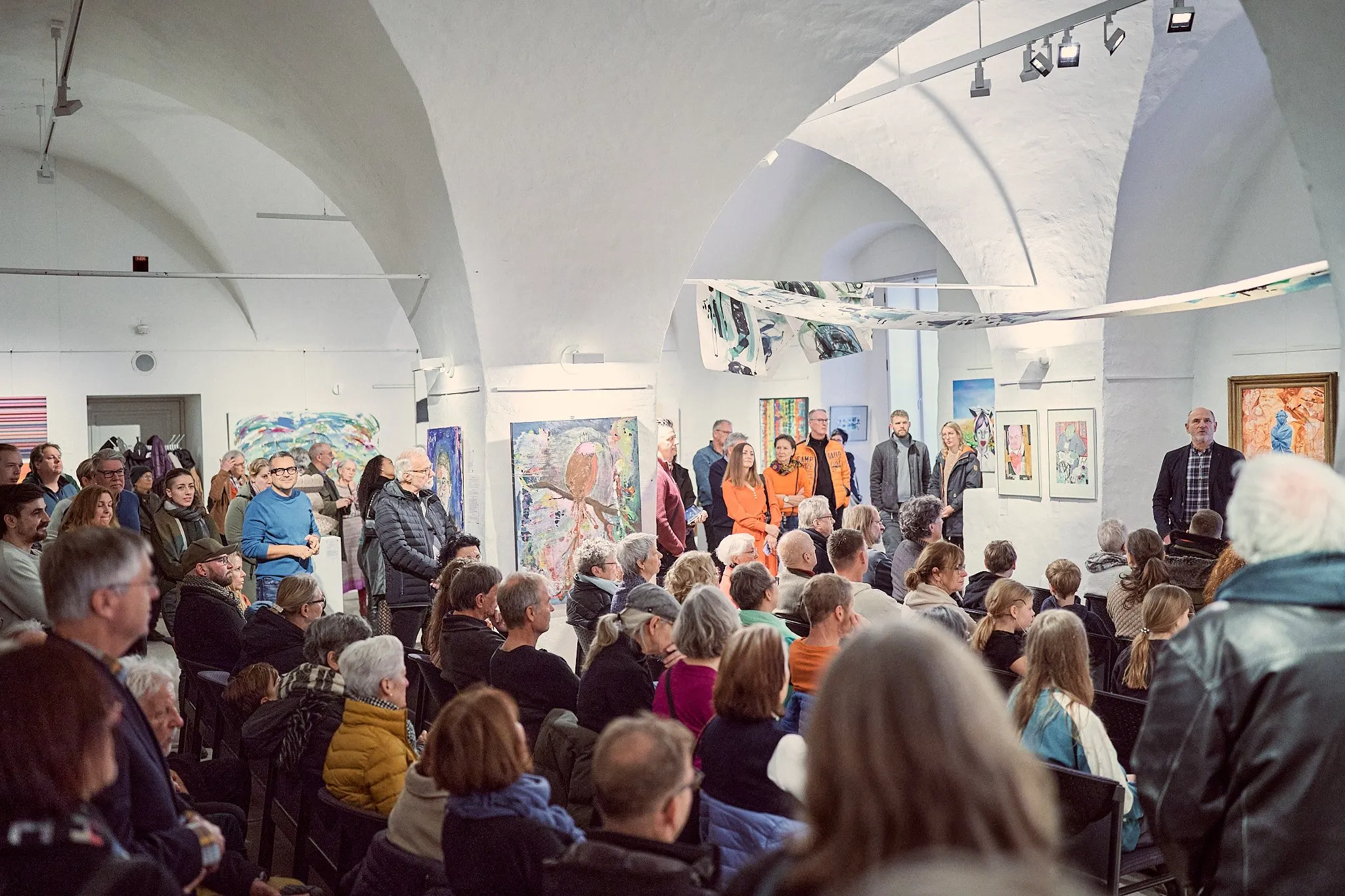 Sitzendes Publikum in der gewölbten Halle bei der Eröffnung, vorne stehen mehrere Personen vor den Werken