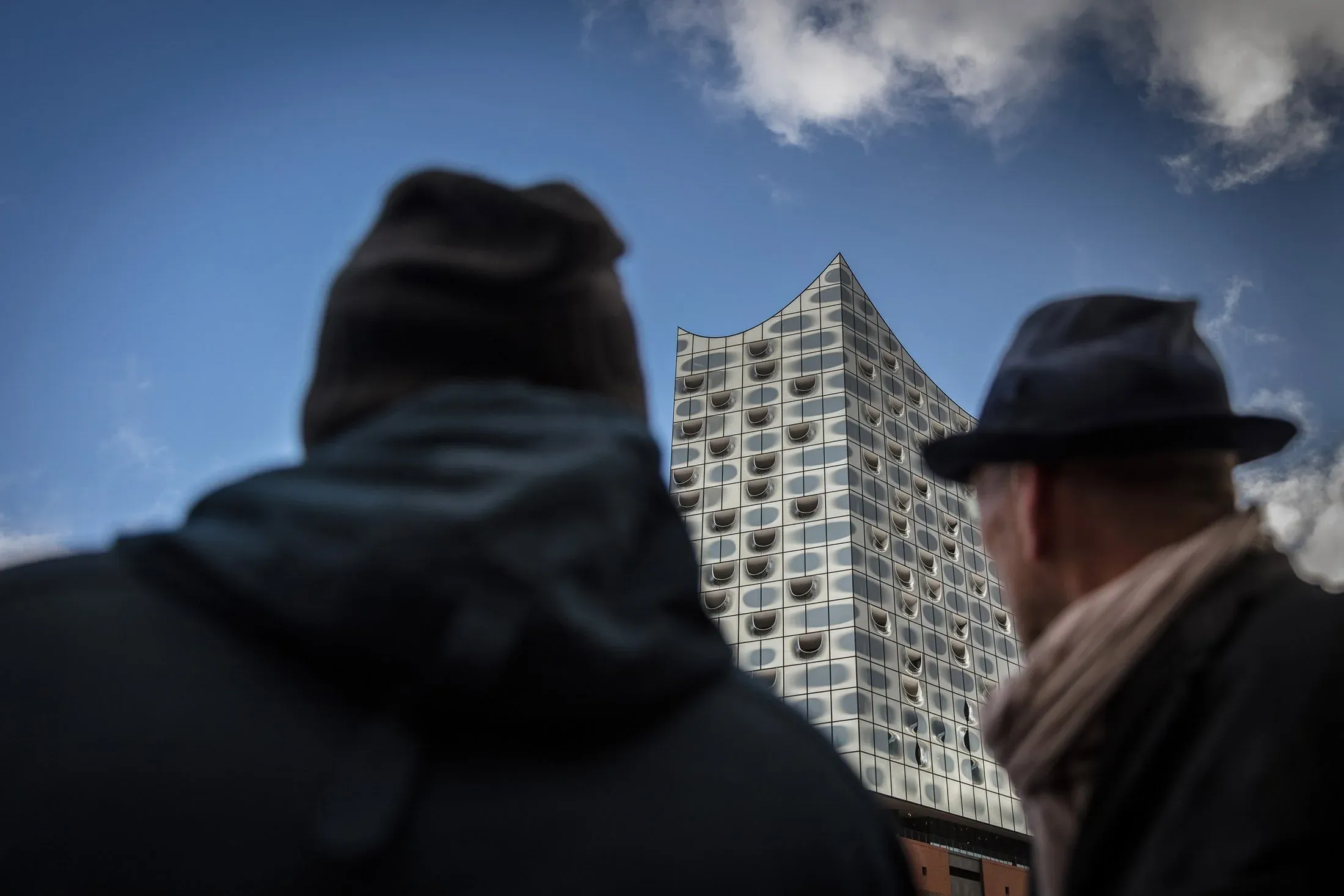 Zwei unscharfe Personen im Vordergrund, dazwischen die Glasfassade der Elbphilharmonie vor blauem Himmel
