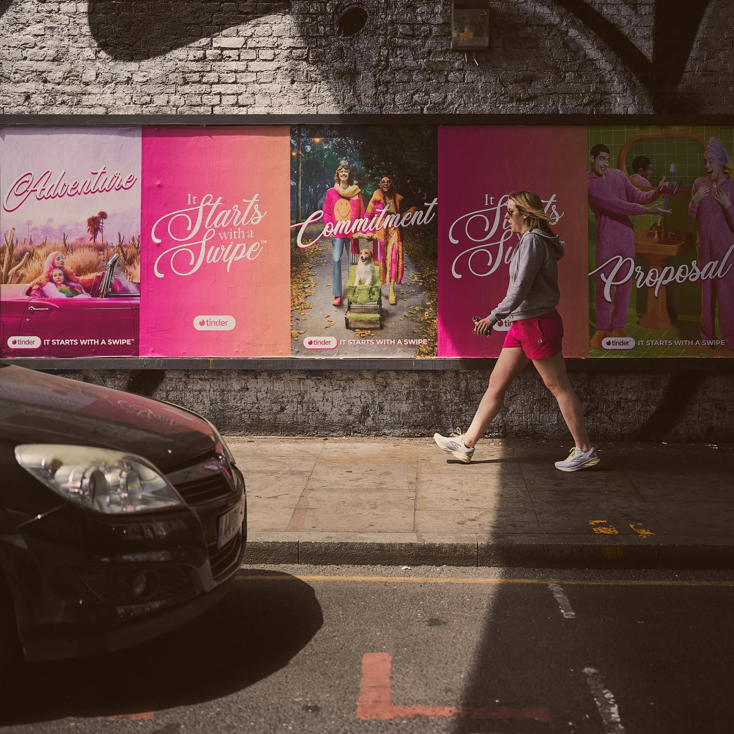 Frau in grauem Pullover joggt an einer Reihe pinker Plakate vorbei, parkendes Auto im Vordergrund
