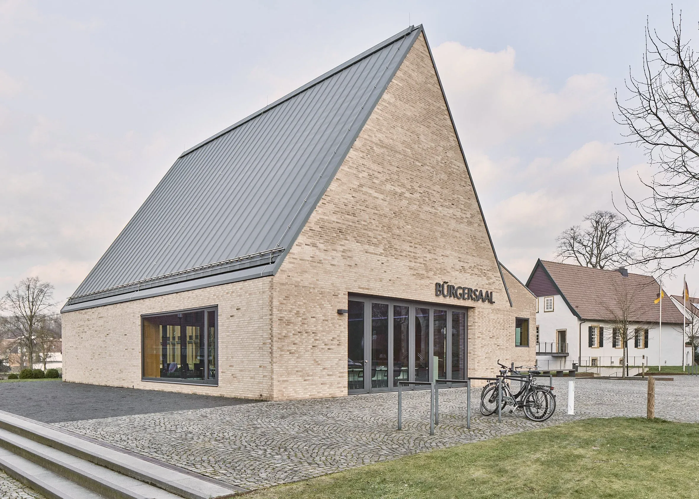 Außenansicht des Bürgersaals mit steilem Metalldach und Schriftzug Bürgersaal, davor abgestelltes Fahrrad und Wohnhäuser