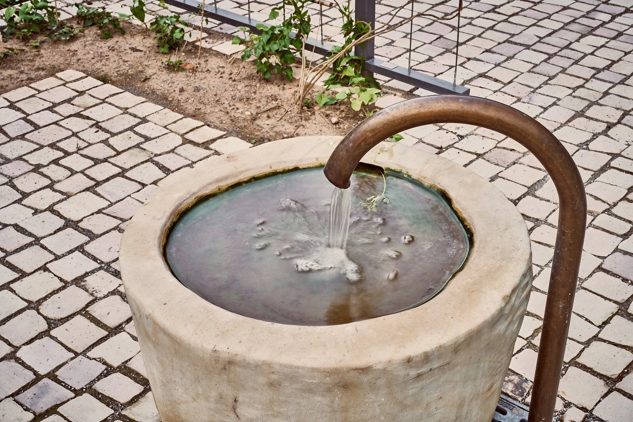 Steinbrunnen mit Wasserzulauf aus einem gebogenen Rohr, umgeben von hellem Kopfsteinpflaster