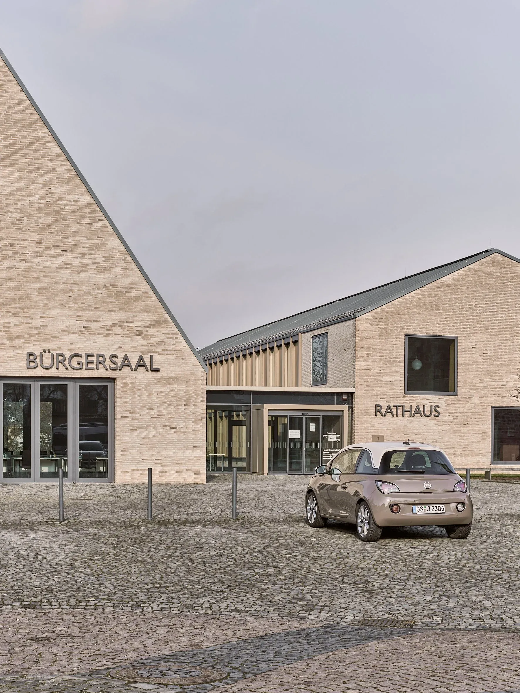 Außenansicht mit den Giebeln von Bürgersaal und Rathaus, gemeinsamem Eingang und einem parkenden Auto auf dem gepflasterten Vorplatz