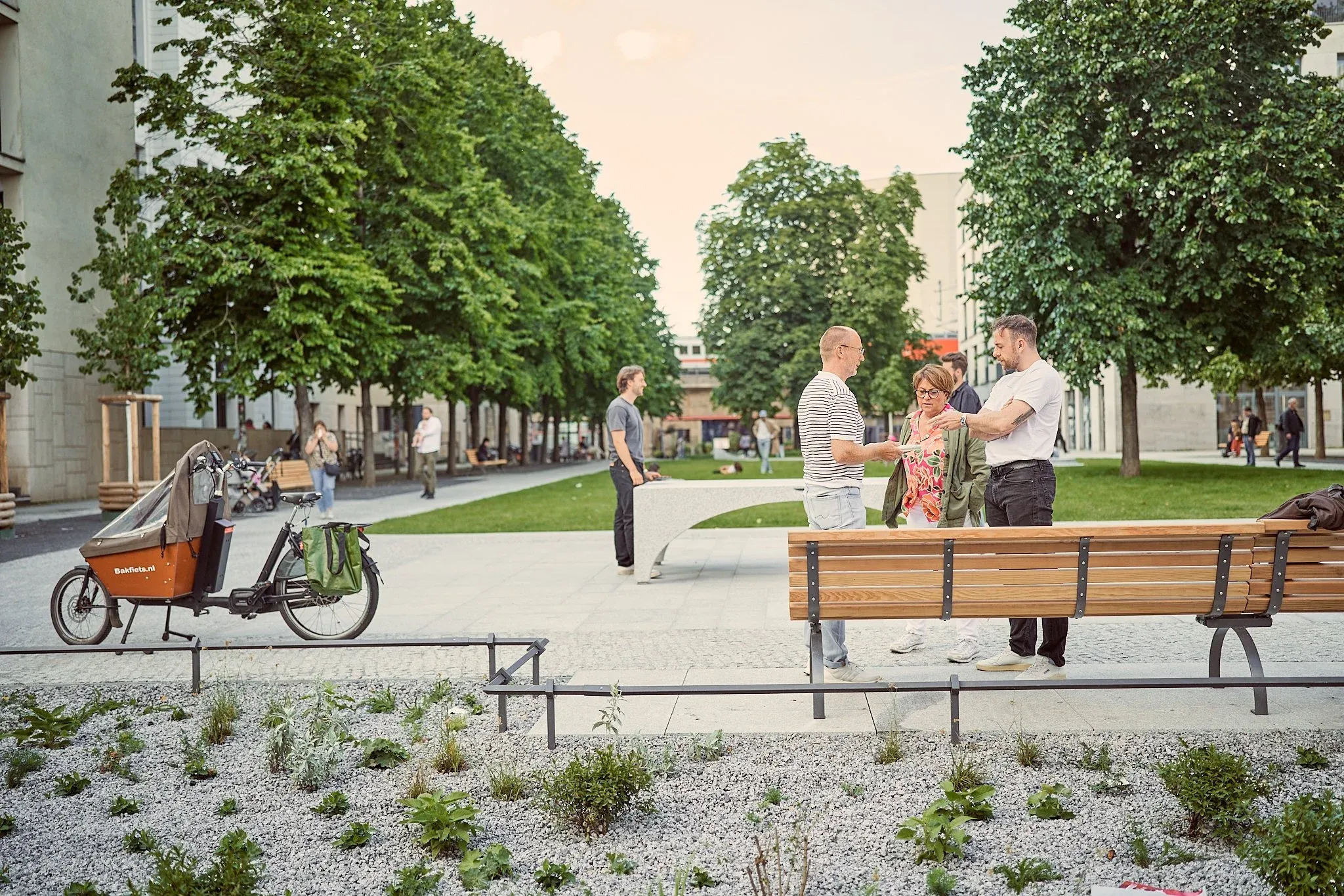 Städtischer Platz mit Bäumen, langen Holzbänken, Lastenrad und stehenden Personen im Abendlicht