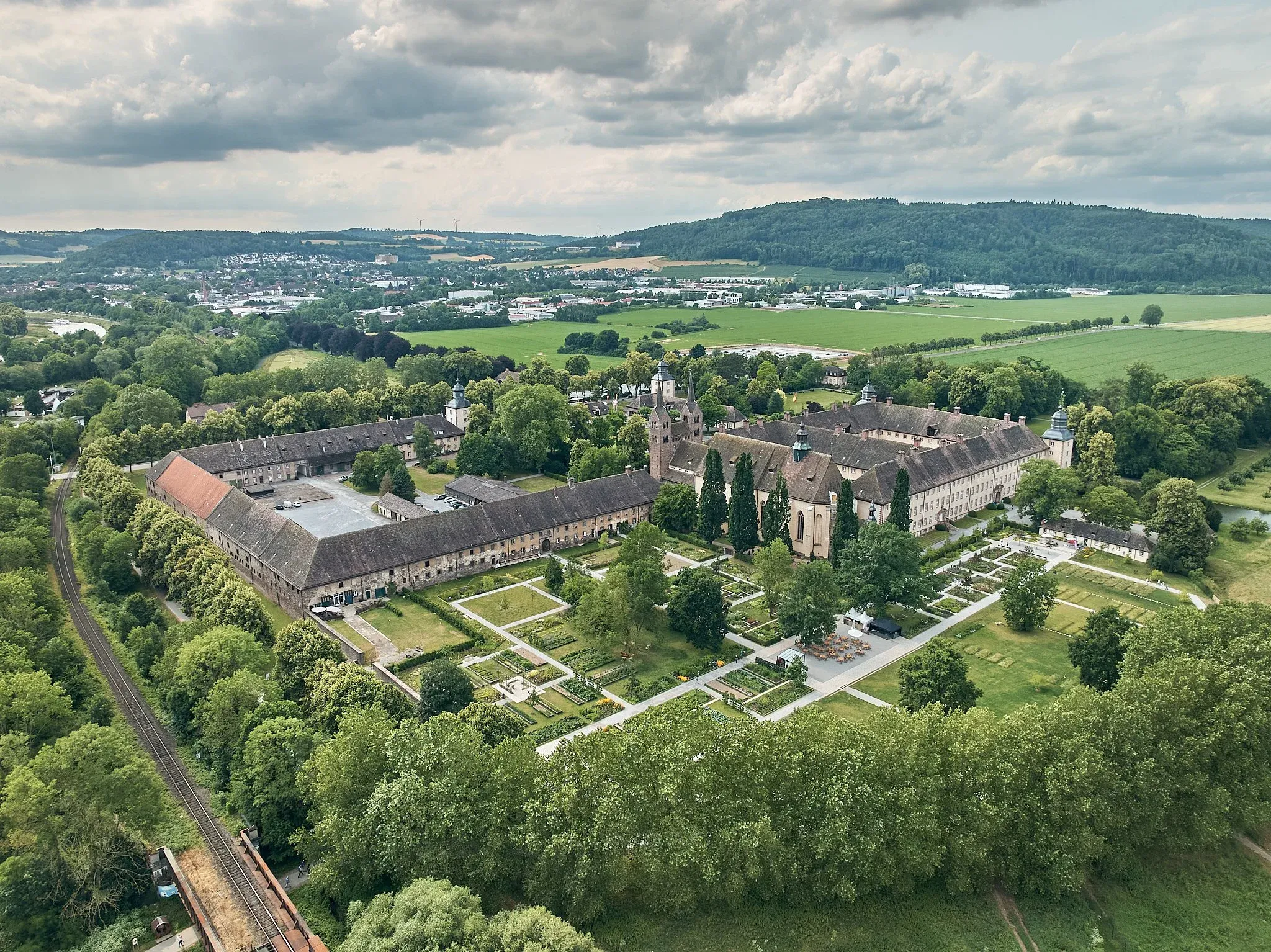 Luftaufnahme der Klosteranlage Corvey in Höxter mit Remtergarten, Wiesen und Bahnlinie unter bewölktem Himmel