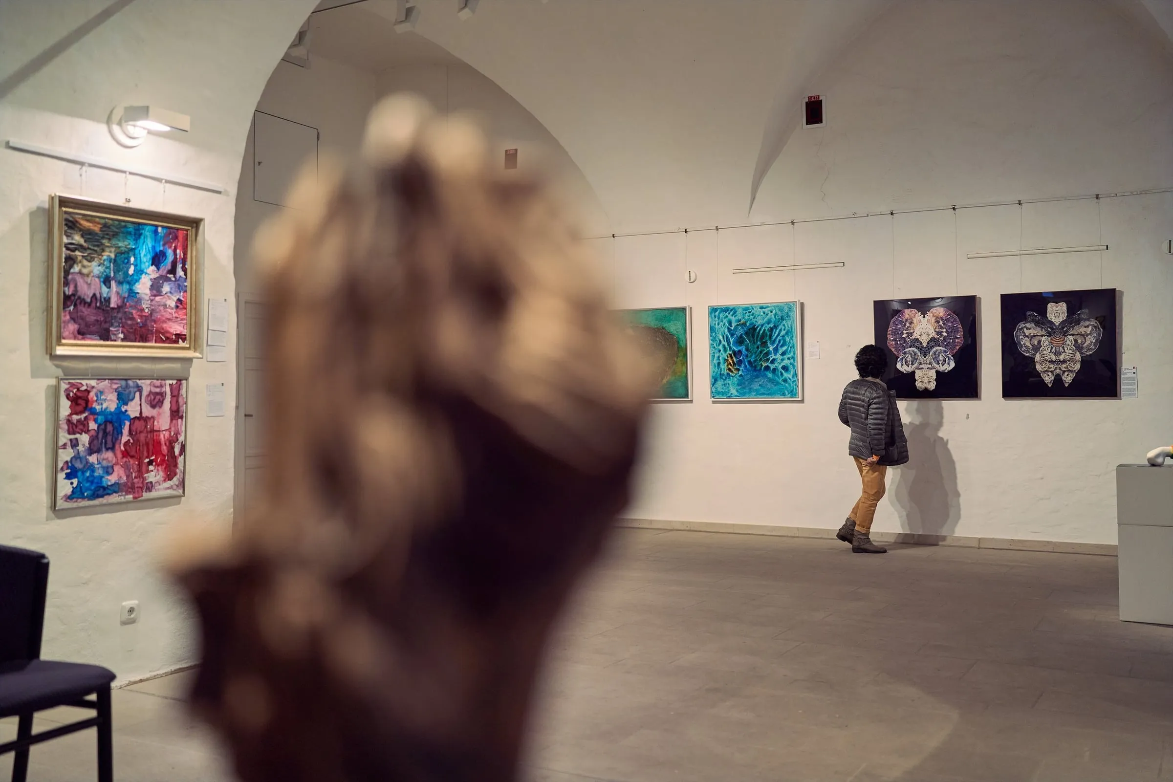 Person betrachtet abstrakte Werke an weißer Wand im gewölbten Ausstellungssaal, vorn unscharf eine Skulptur