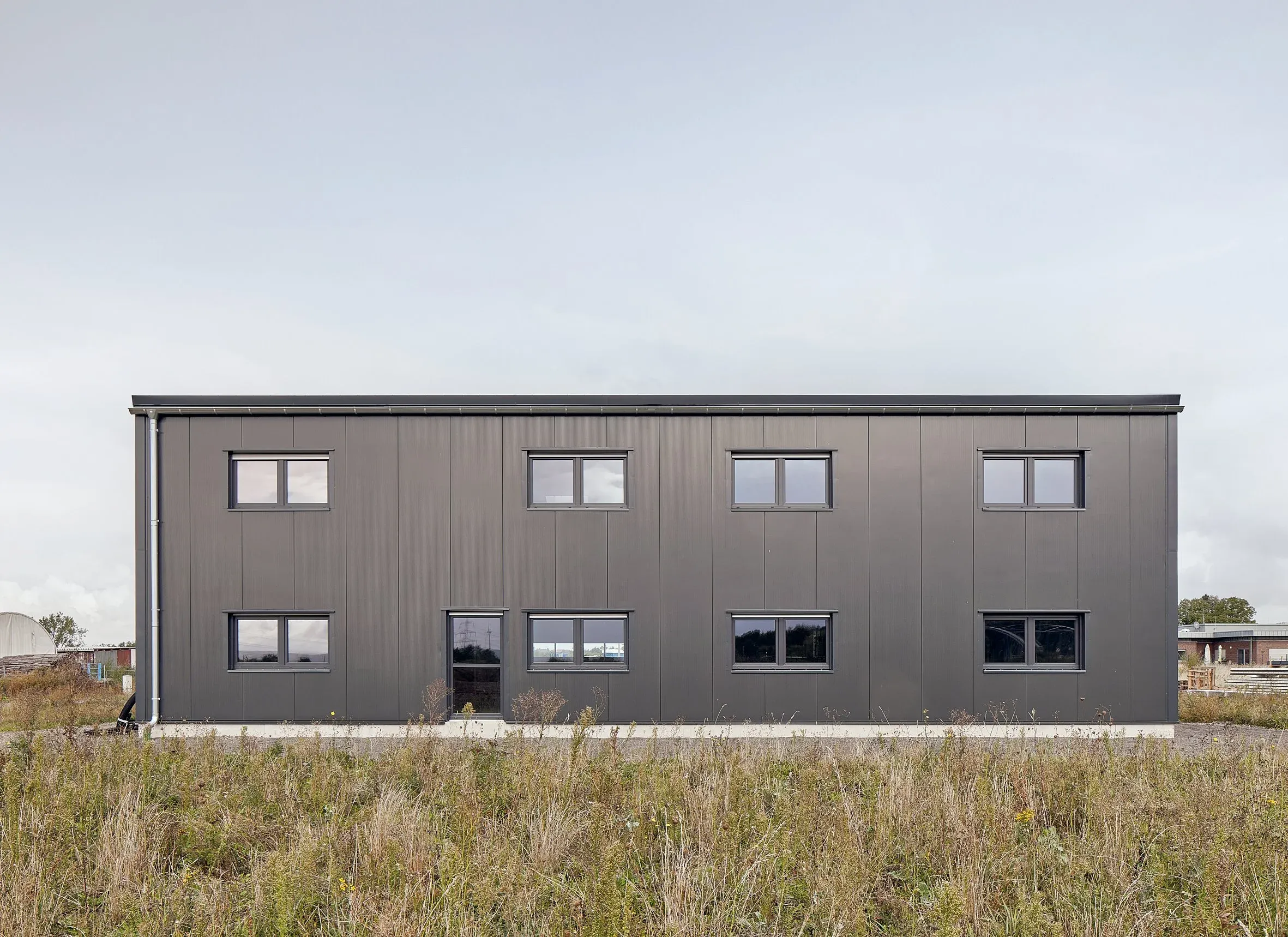 Außenansicht des grauen zweigeschossigen HTS-Funktionsbaus mit Stehfalzfassade und Fensterreihen, davor Wiese