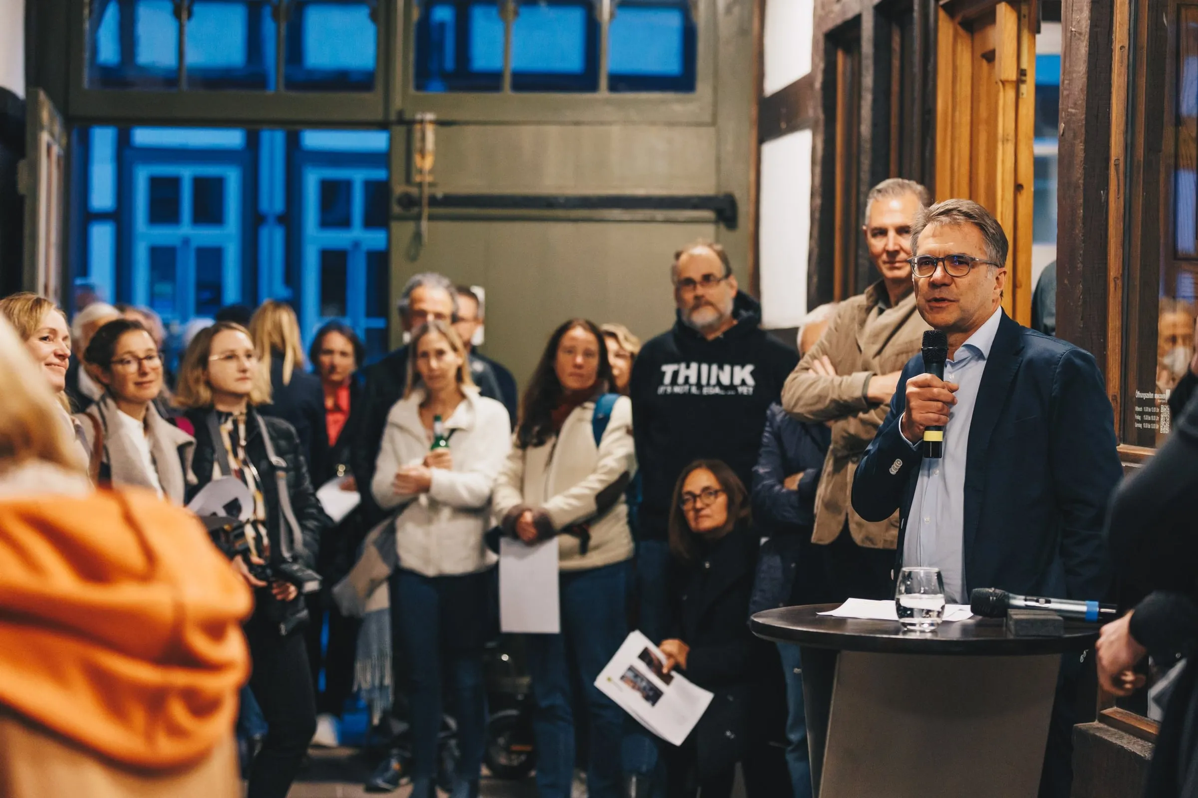 Mann mit Mikrofon spricht zu einer Gruppe von Gästen in einem holzvertäfelten Raum