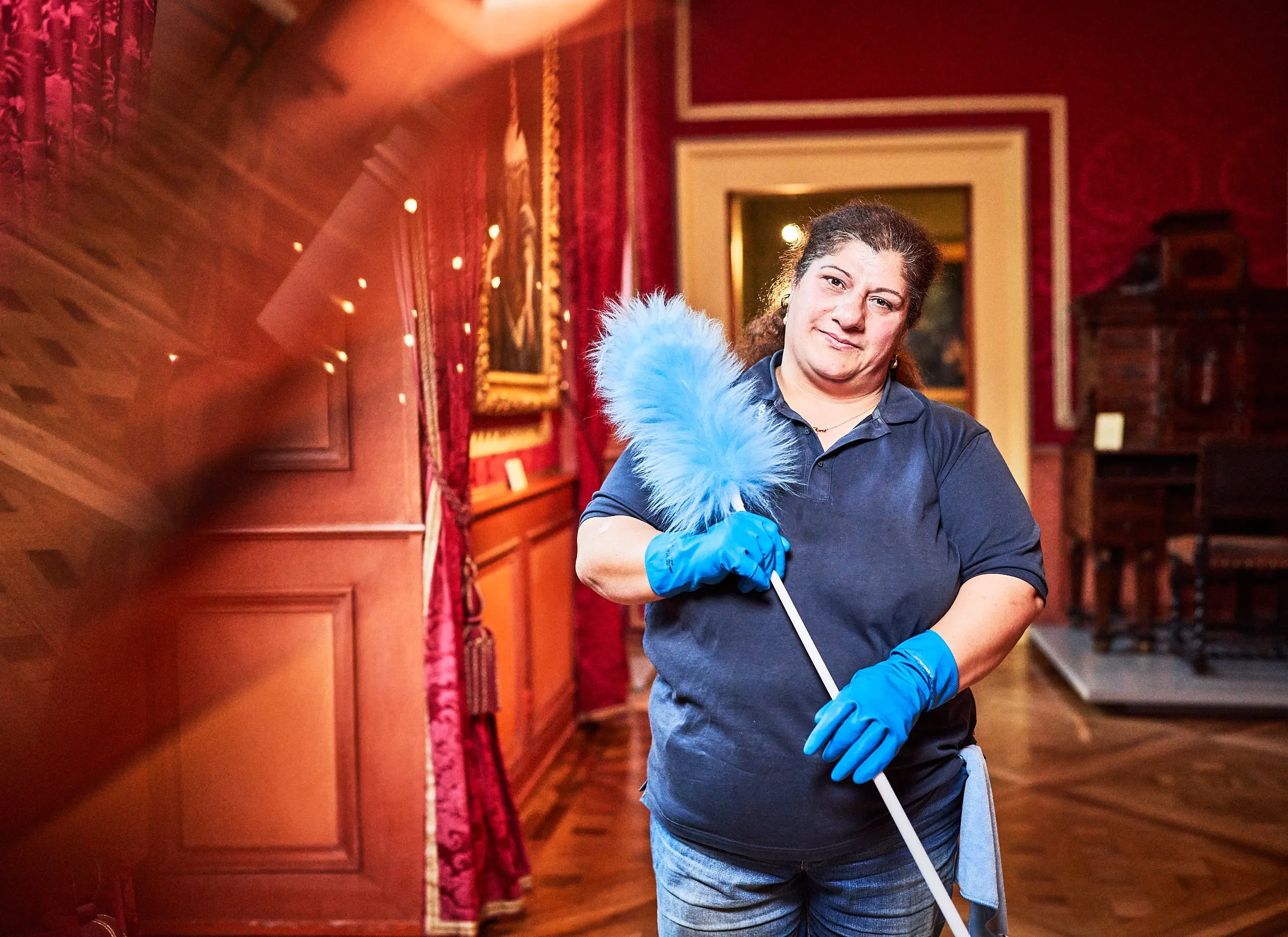 Porträt einer lächelnden Mitarbeiterin mit hellblauem Staubwedel im roten Schlosssaal, Innenaufnahme