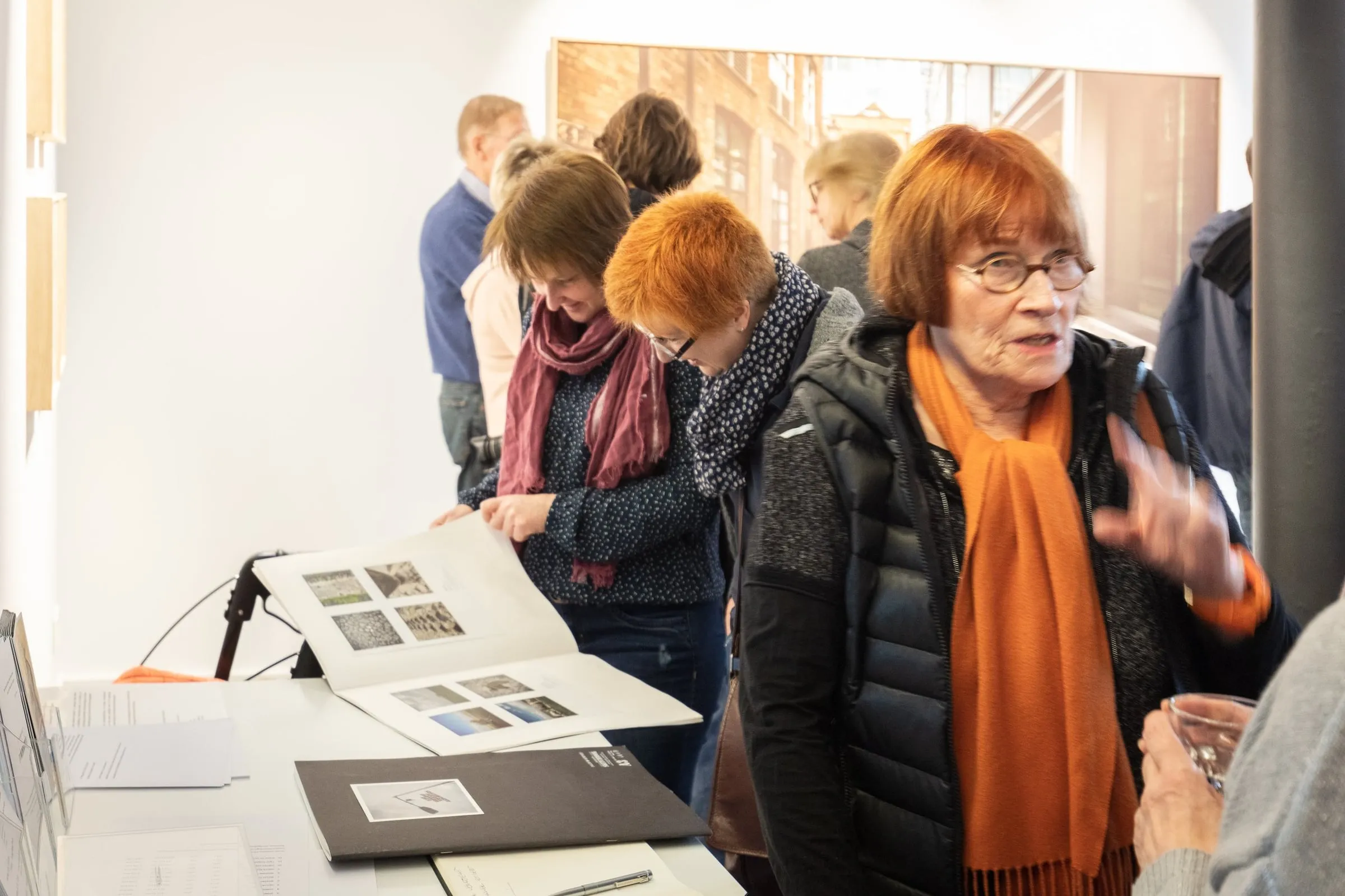Besucherinnen blättern an einem Tisch in einem Bildband, im Hintergrund weitere Gäste