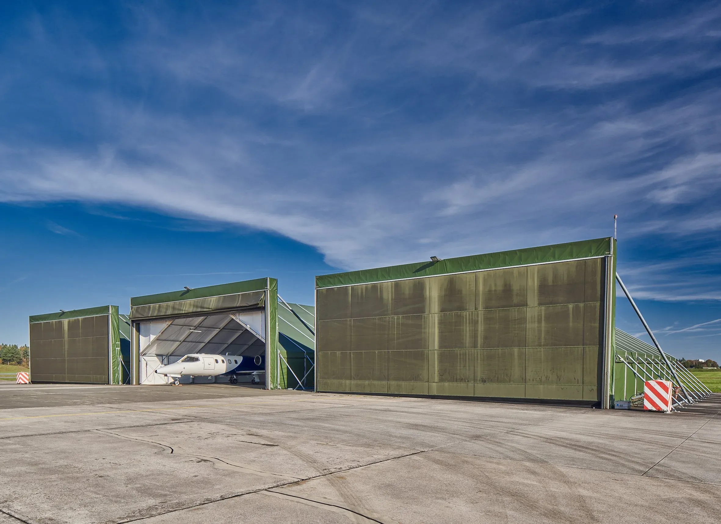 Außenaufnahme, olivgrüne Hallen über einem Flugzeughangar mit Geschäftsjet auf dem Flugfeld, blauer Himmel