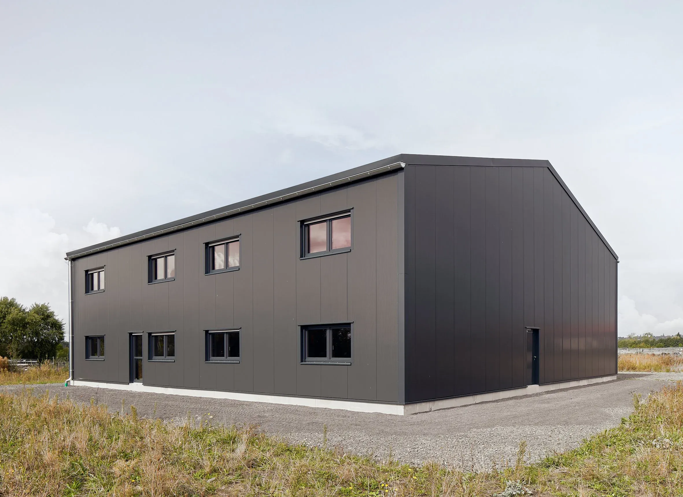 Schräge Außenansicht des grauen HTS-Baus über Eck mit Stehfalzfassade, Fenstern und Zufahrt, bewölkter Himmel