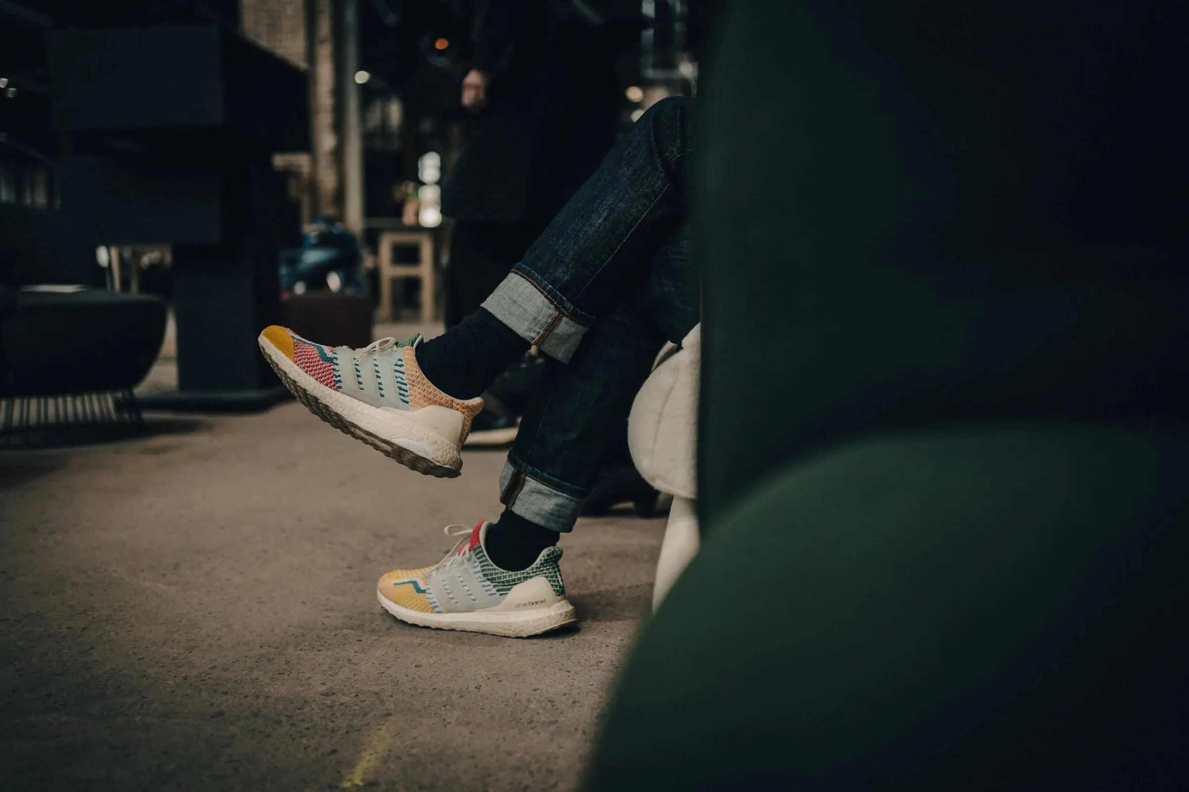 Detail bunter Sneaker einer sitzenden Person mit hochgekrempelter Jeans in dunkler Halle