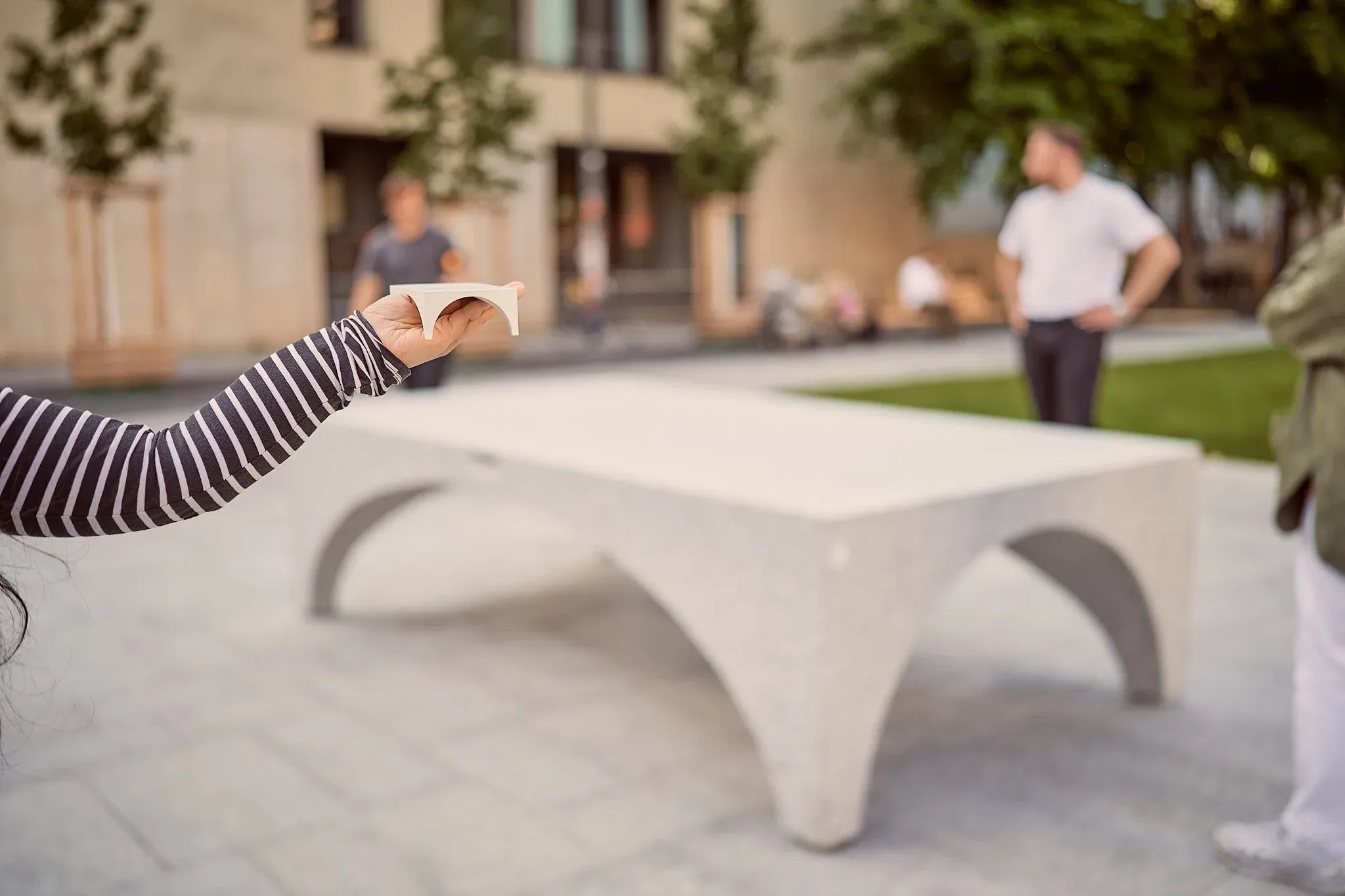 Hand legt ein Papiermodell einer Bank auf den hellen Steintisch mit Bogenfüßen, Personen im Hintergrund