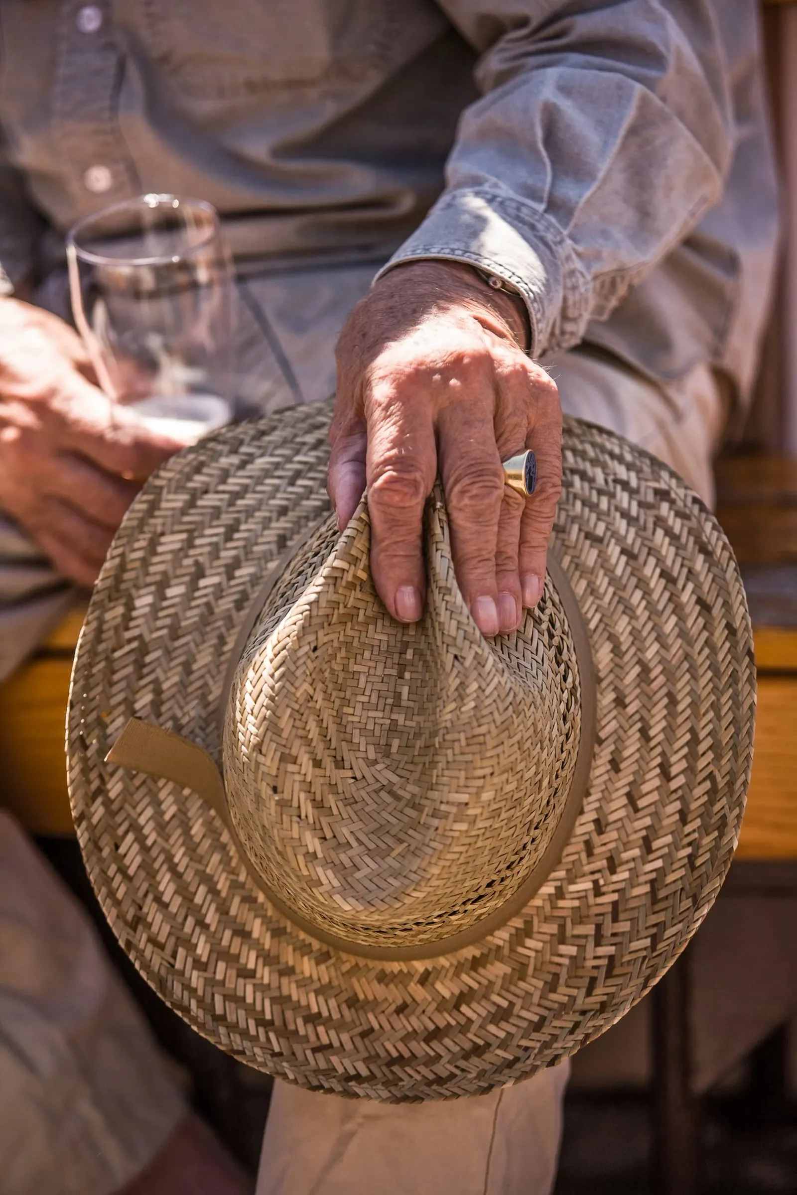 Detailaufnahme einer Hand auf einem Strohhut, im Hintergrund ein sitzender Mann mit Glas, helles Tageslicht
