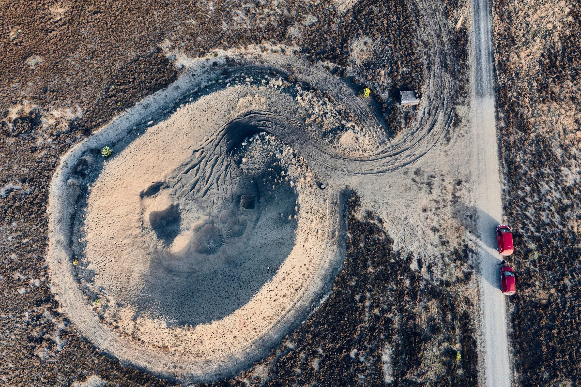 Luftaufnahme einer ausgehobenen Sandmulde neben einer Straße, ein rotes Fahrzeug fährt vorbei