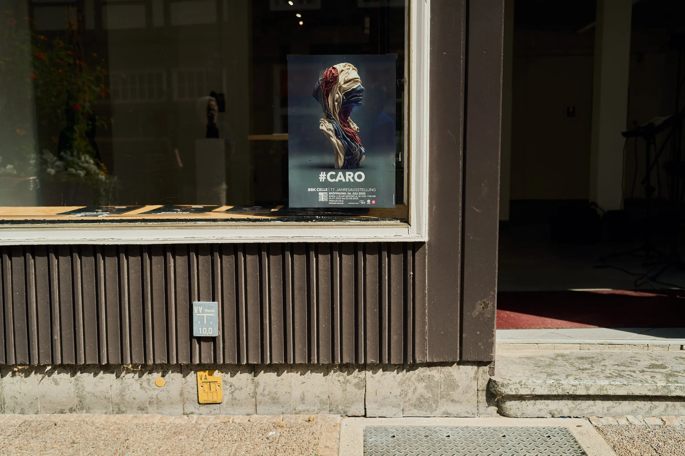 Schaufenster mit dem Plakat zur Ausstellung #CARO, davor die Hausfassade in der Sonne