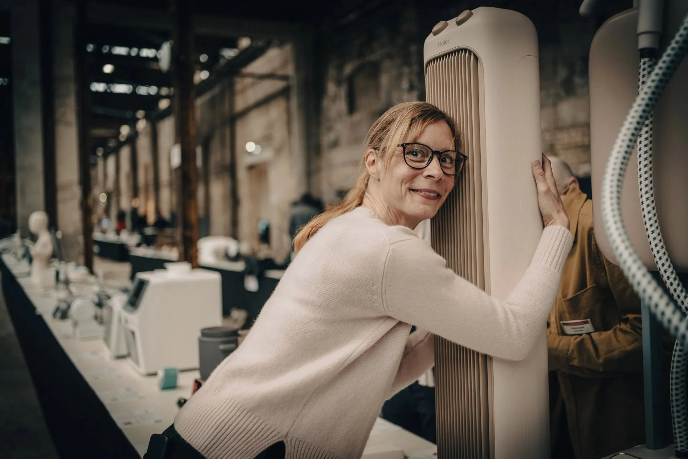 Lächelnde Frau mit Brille hält ein längliches Objekt am Ausstellungstisch, weitere Exponate dahinter