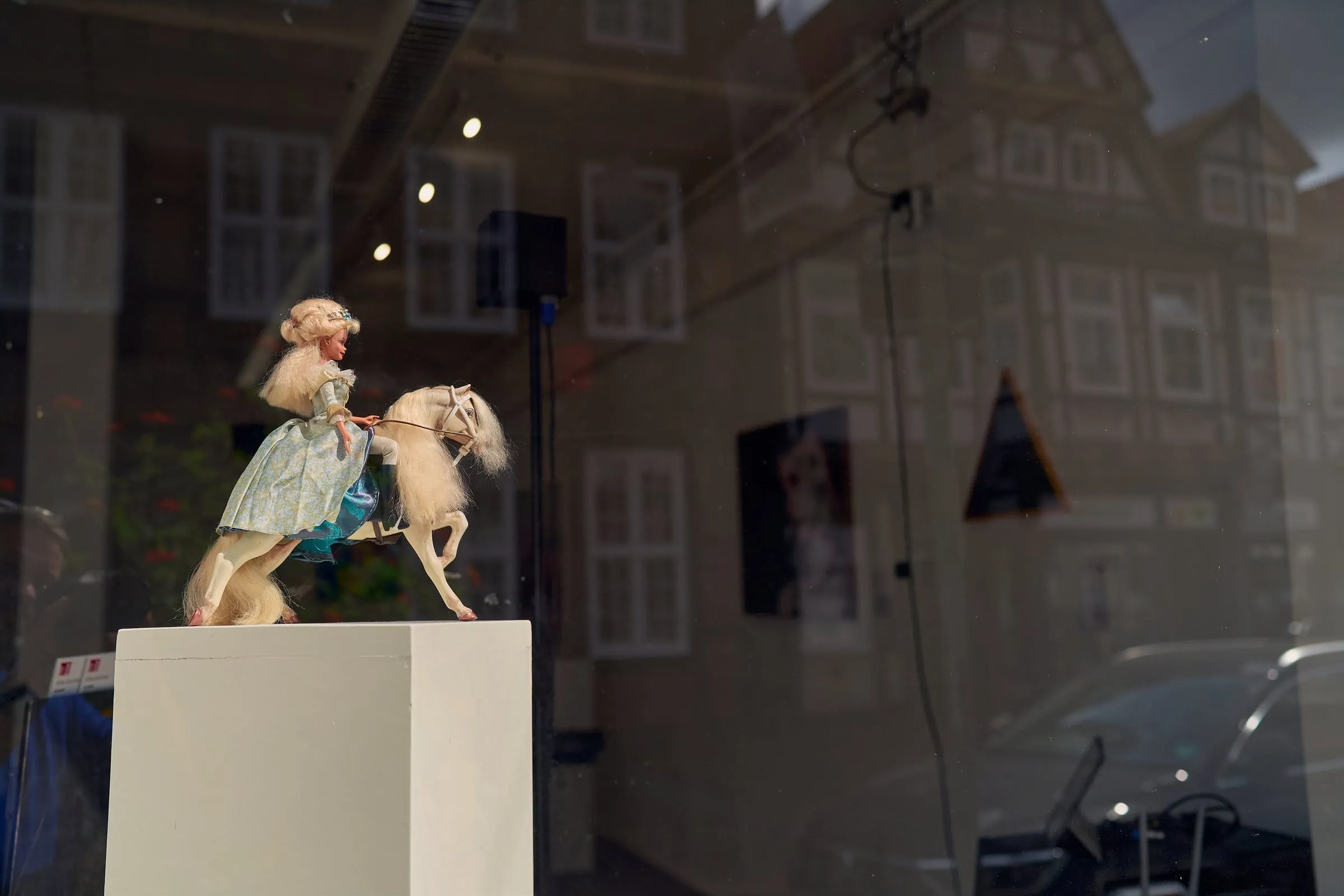 Eine Puppe auf einem Pferd als Objekt auf einem Sockel im Schaufenster, im Glas spiegelt sich die Fachwerkstraße