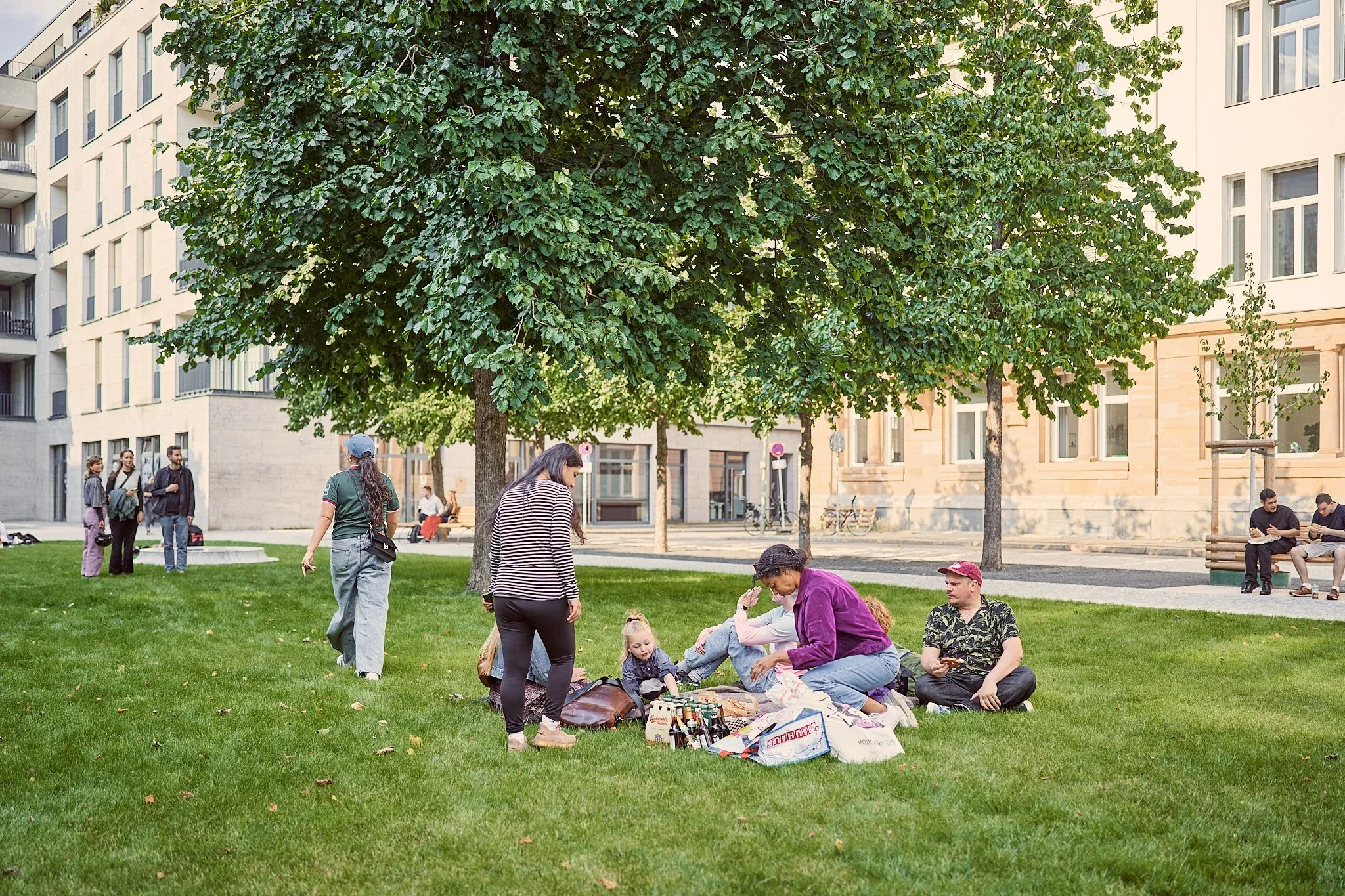 Menschen sitzen auf einer Picknickdecke im Schatten eines Baumes auf dem Rasen, Gebäude dahinter