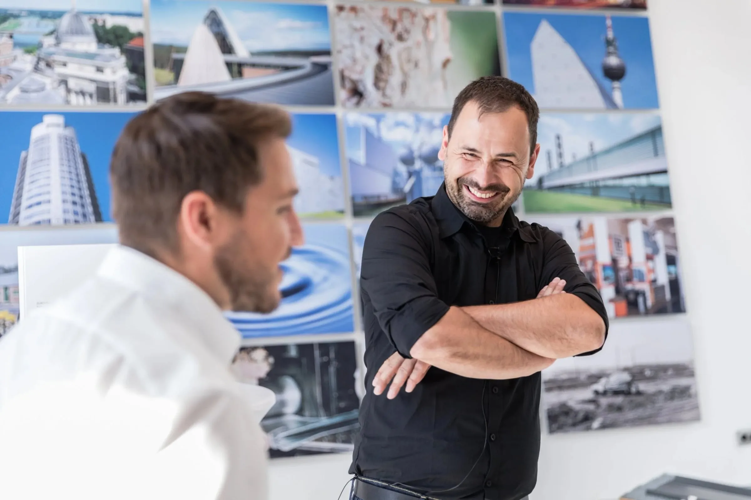 Lachender Mann in schwarzem Hemd mit verschränkten Armen im Gespräch vor einer Wand mit Architekturfotografien