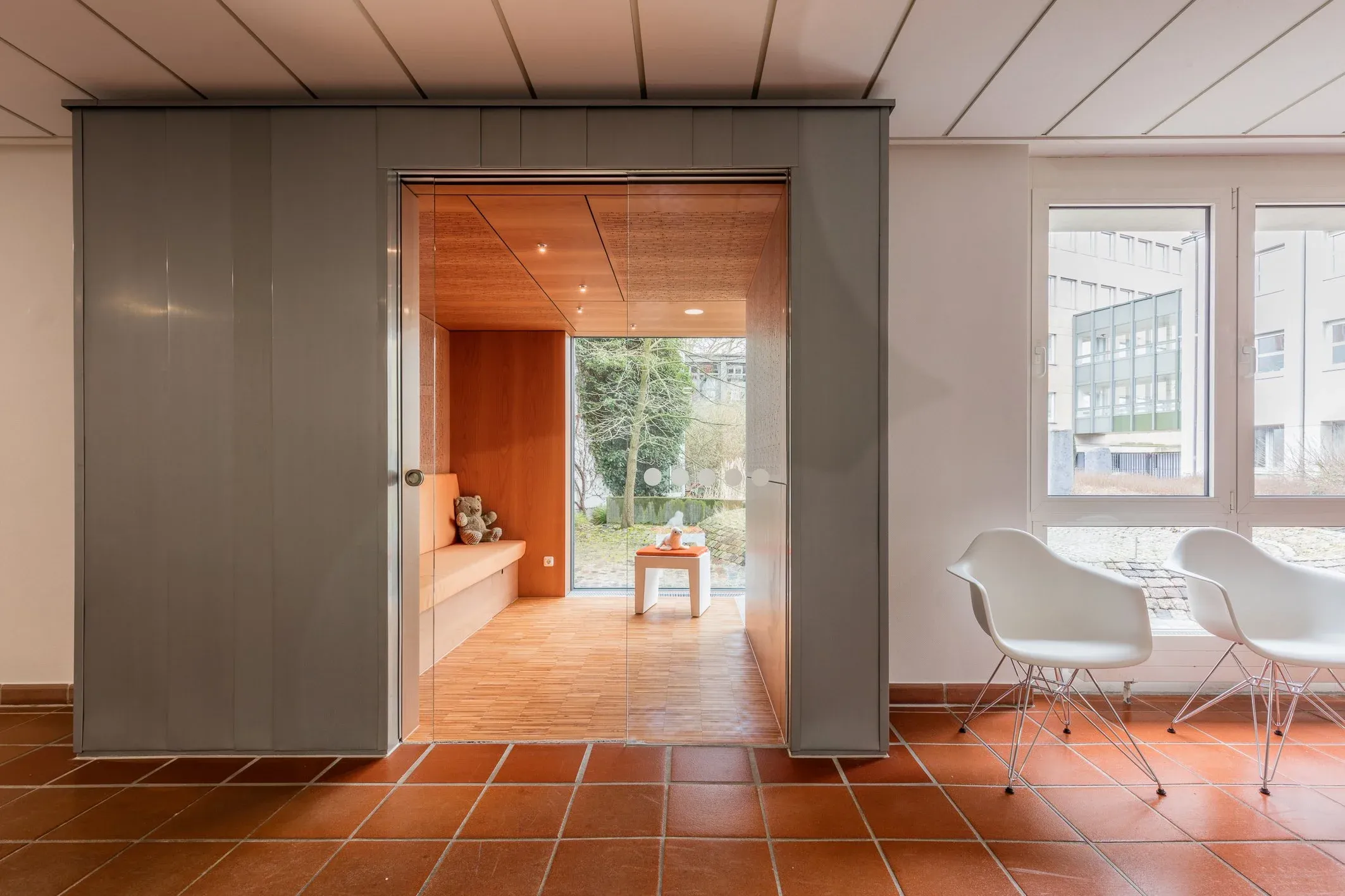 Innenraum mit grauer Schiebewand, dahinter Sitznische aus Holz, Fliesenboden und weiße Stühle am Fenster