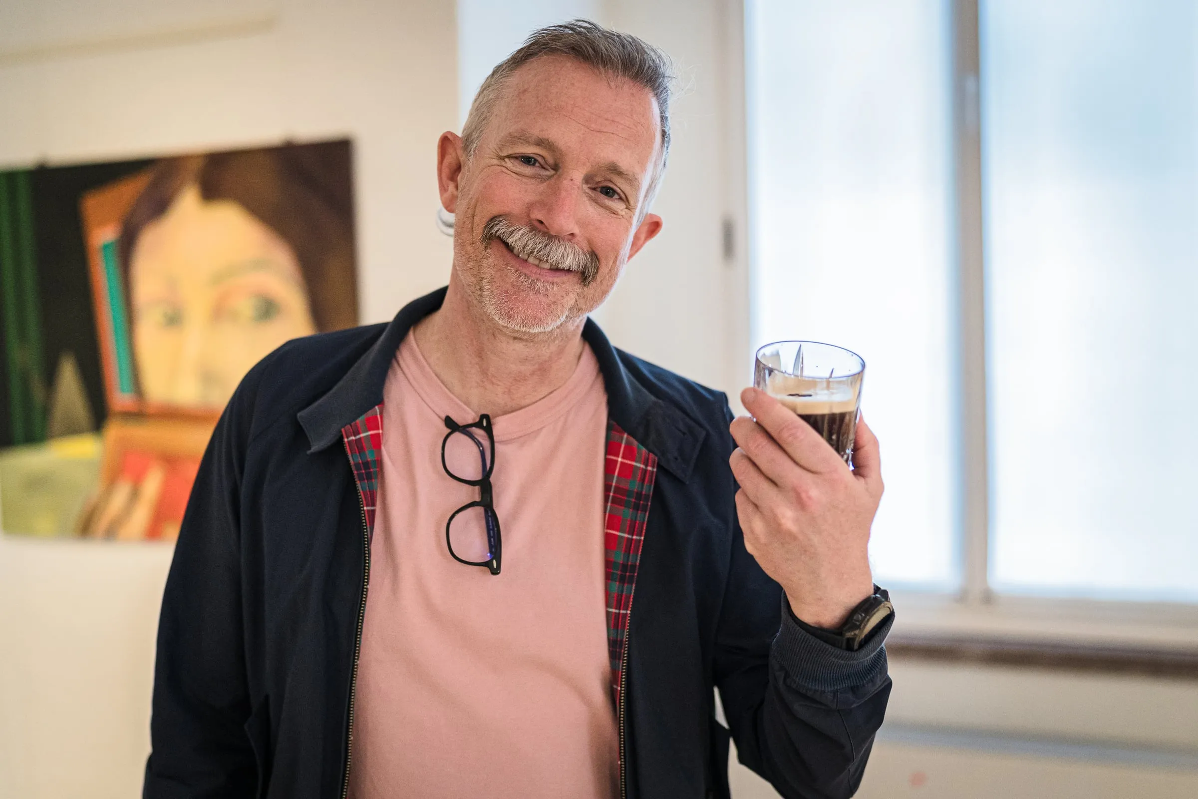 Lächelnder Mann mit Getränk im Galerieraum, im Hintergrund ein gerahmtes Werk und ein Fenster