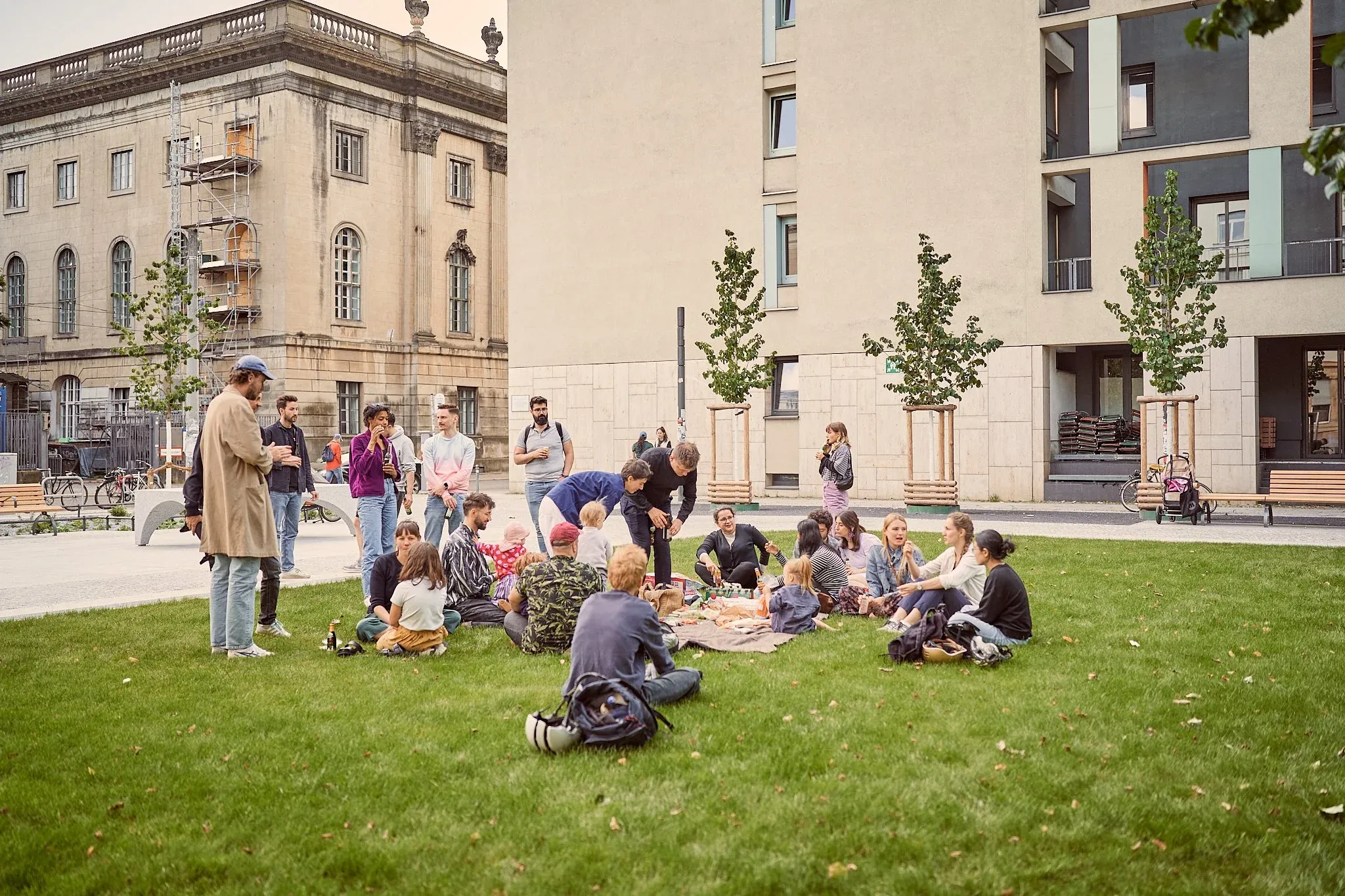 Größere Gruppe sitzt im Kreis auf dem Rasen, Personen stehen daneben, Gebäude und Bäume rundum