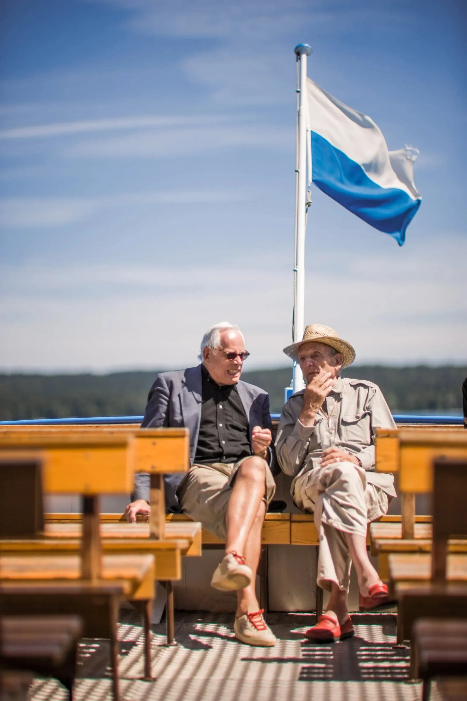 Zwei Männer auf einem Schiff sitzend im Gespräch, über ihnen eine wehende blauweiße Flagge, blauer Himmel