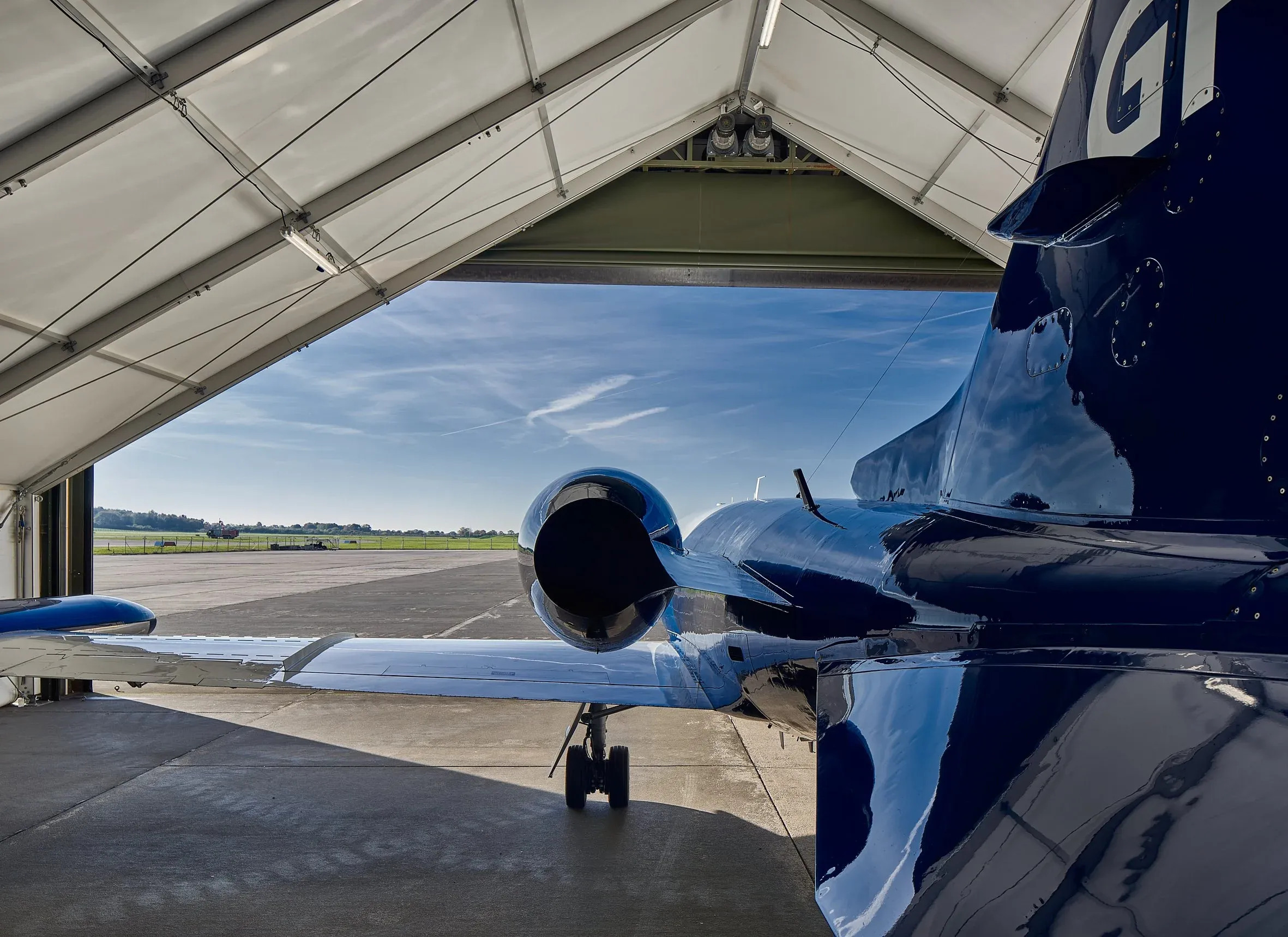 Detail im Hangar, Heck und Triebwerk eines blauen Geschäftsjets unter heller Zelthülle, Blick aufs Flugfeld