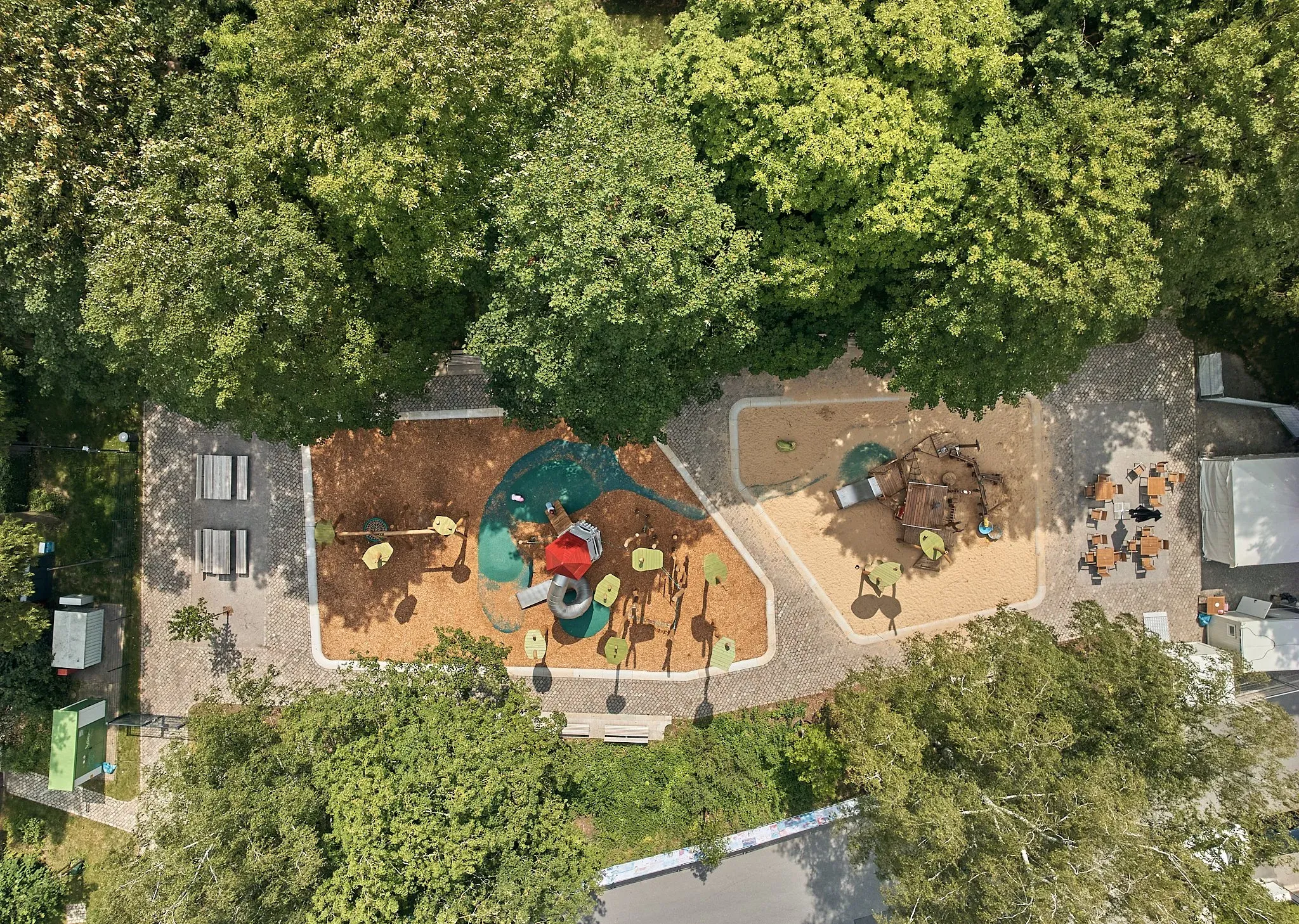 Luftaufnahme eines Spielplatzes im Park mit Sandflächen und Spielgeräten, dicht von grünen Baumkronen umrahmt