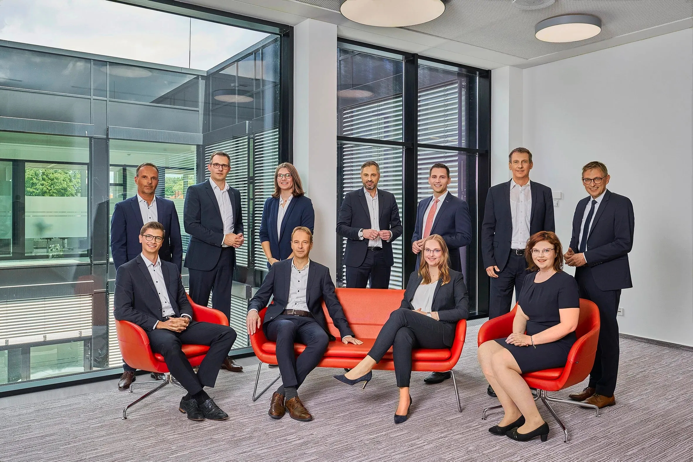 Elfköpfiges Team in Geschäftskleidung in einem Büro, vier Personen sitzen auf roten Sesseln, sieben stehen dahinter