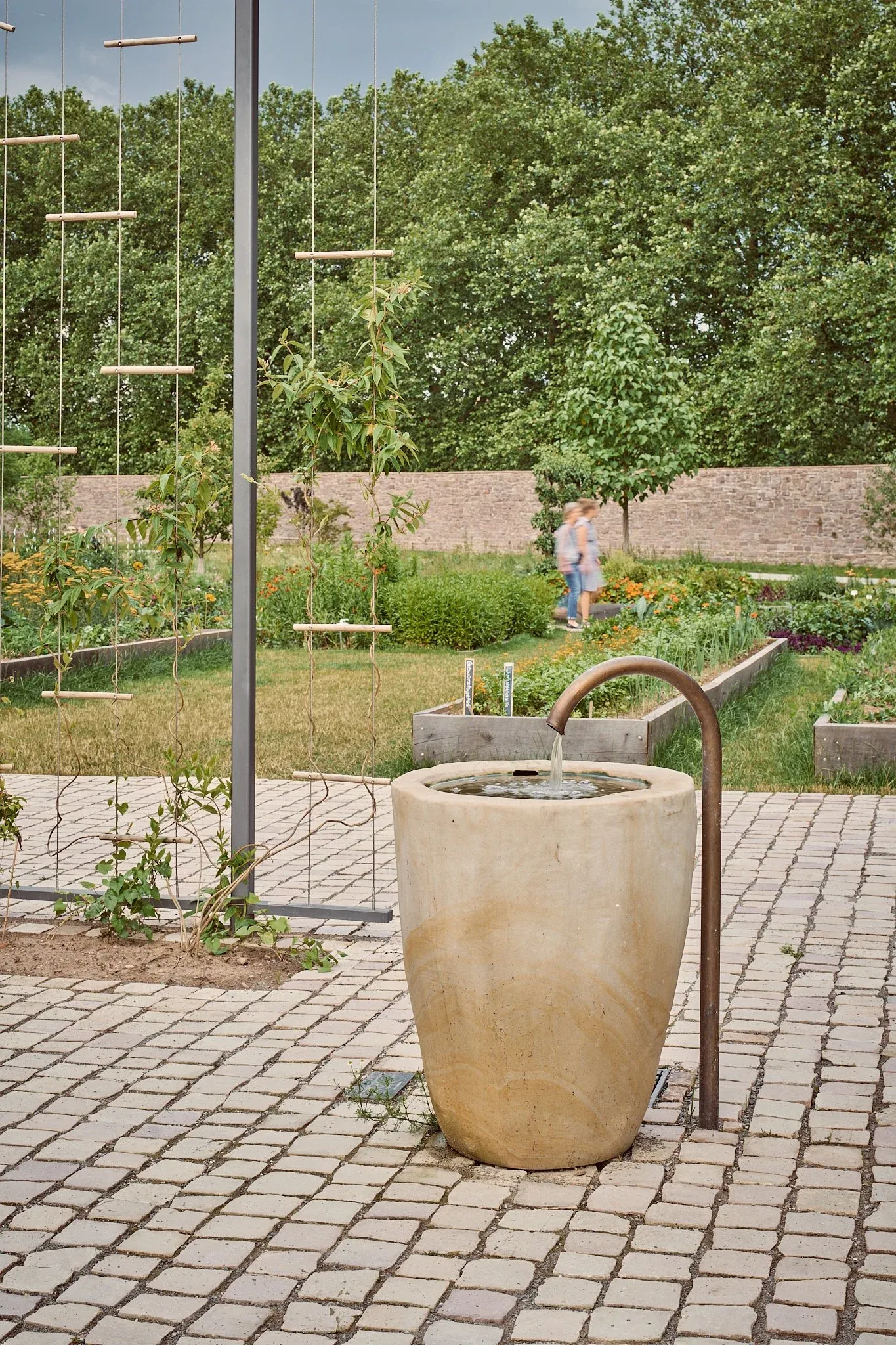 Steinbrunnen mit Wasserzulauf auf gepflastertem Platz im Remtergarten, Beete, Mauer und Bäume im Hintergrund