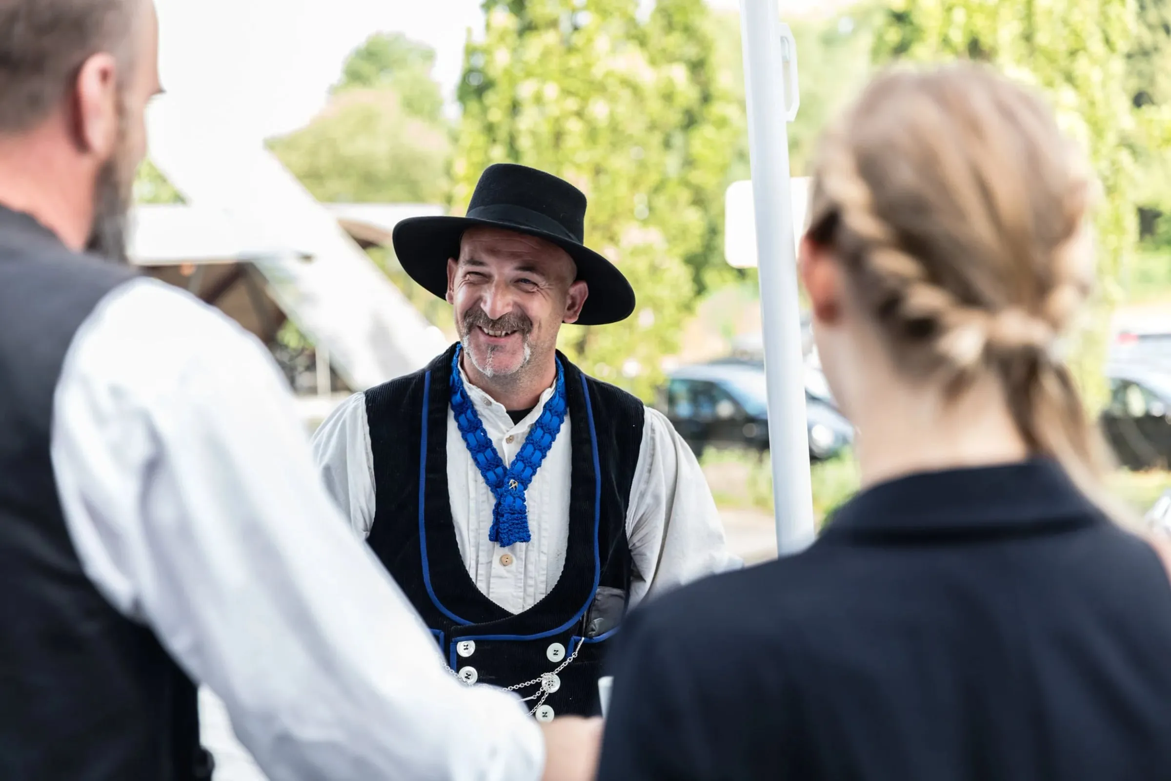 Lachender Mann mit Hut und Trachtenweste im Freien im Gespräch mit zwei Personen, im Hintergrund Bäume und Parkplatz