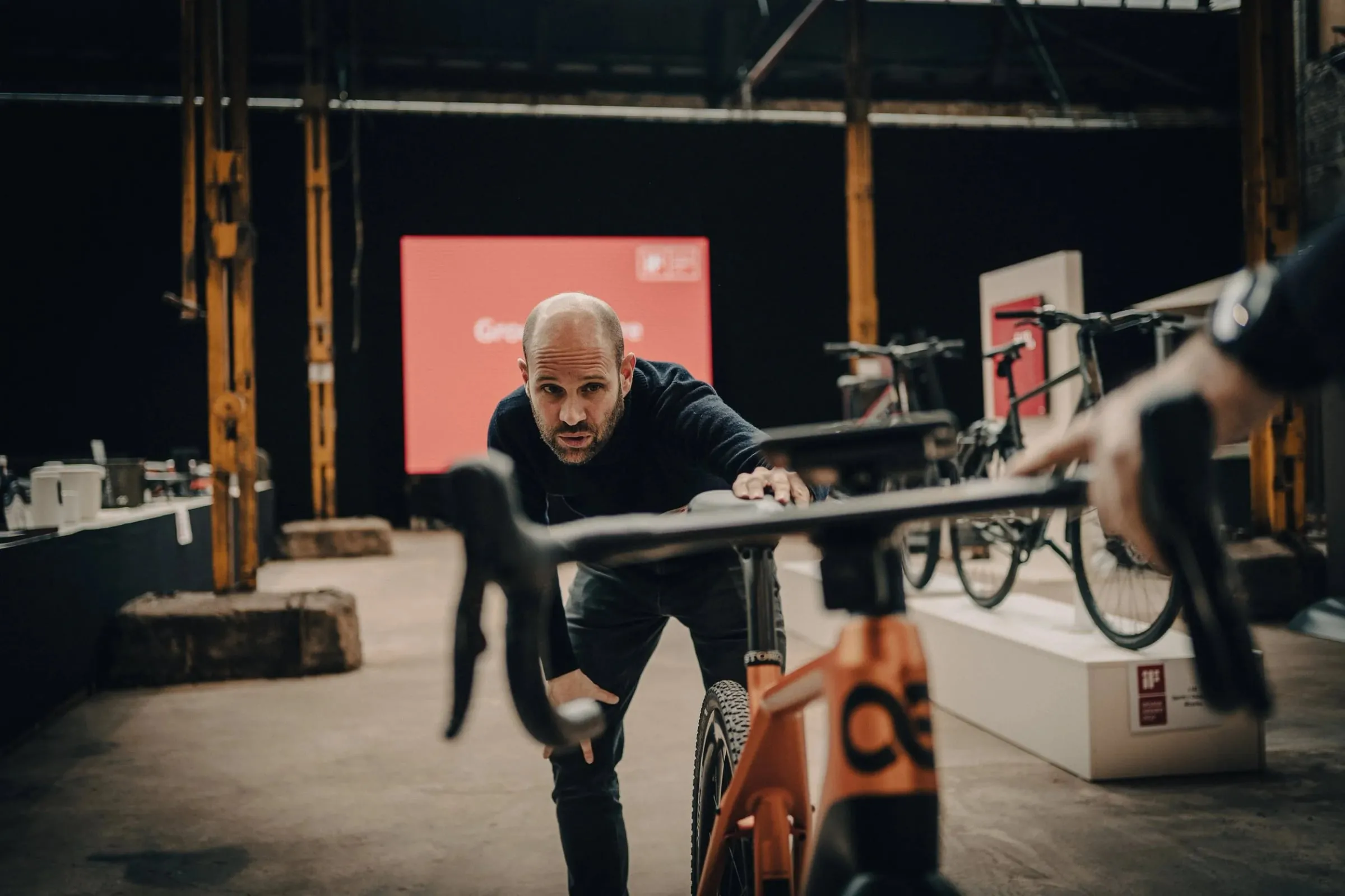Mann beugt sich über ein orangefarbenes Rennrad in der Halle, rote Bühnenwand und Fahrräder dahinter