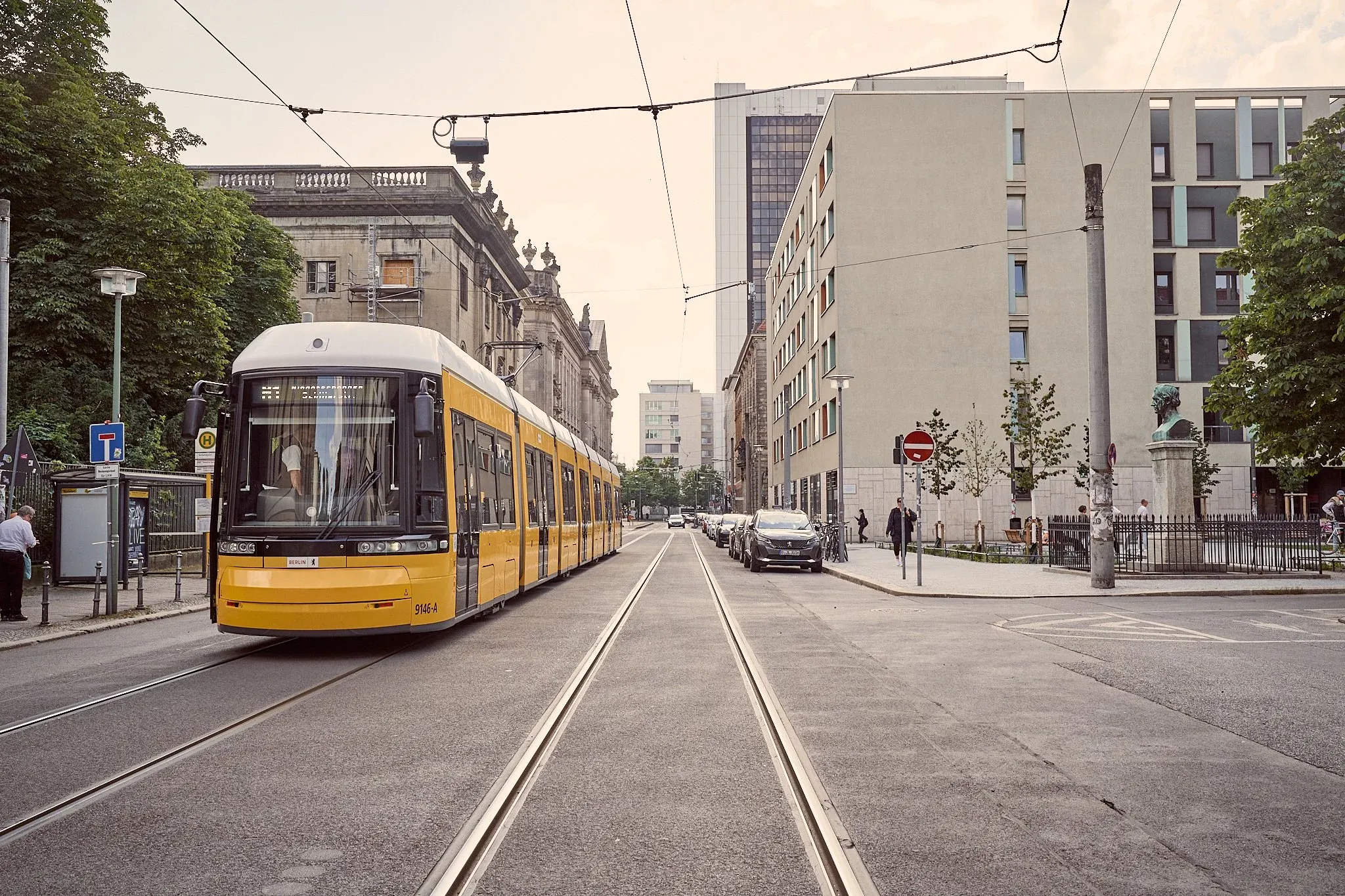 Gelbe Straßenbahn auf Schienen in einer Straße neben dem Platz mit Gründerzeitbau und Neubauten