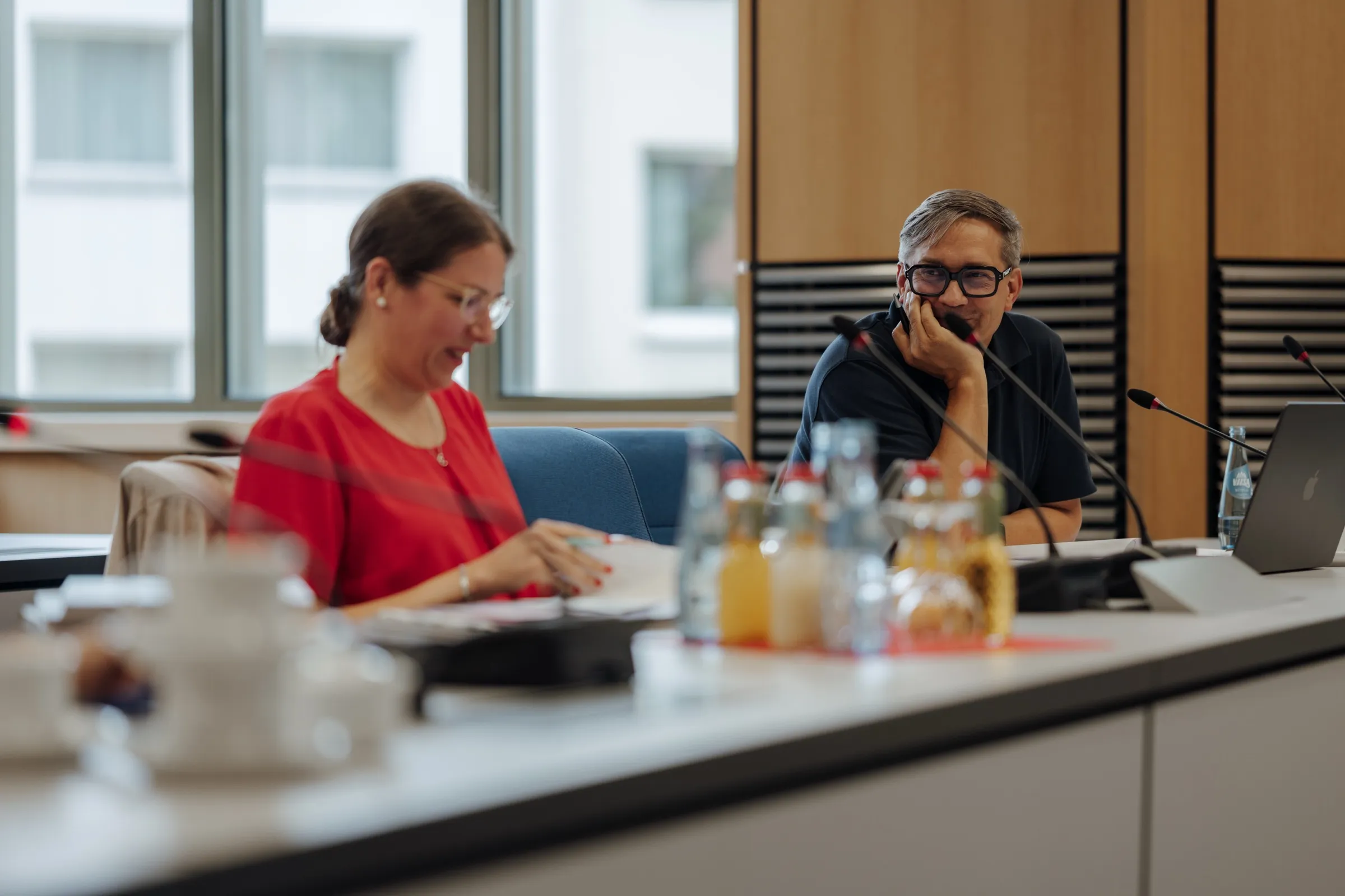 Dr. Anna Heinze and Roman Thomas at the long session table, microphones, water and juice bottles in front of them, tall windows and wooden walls behind