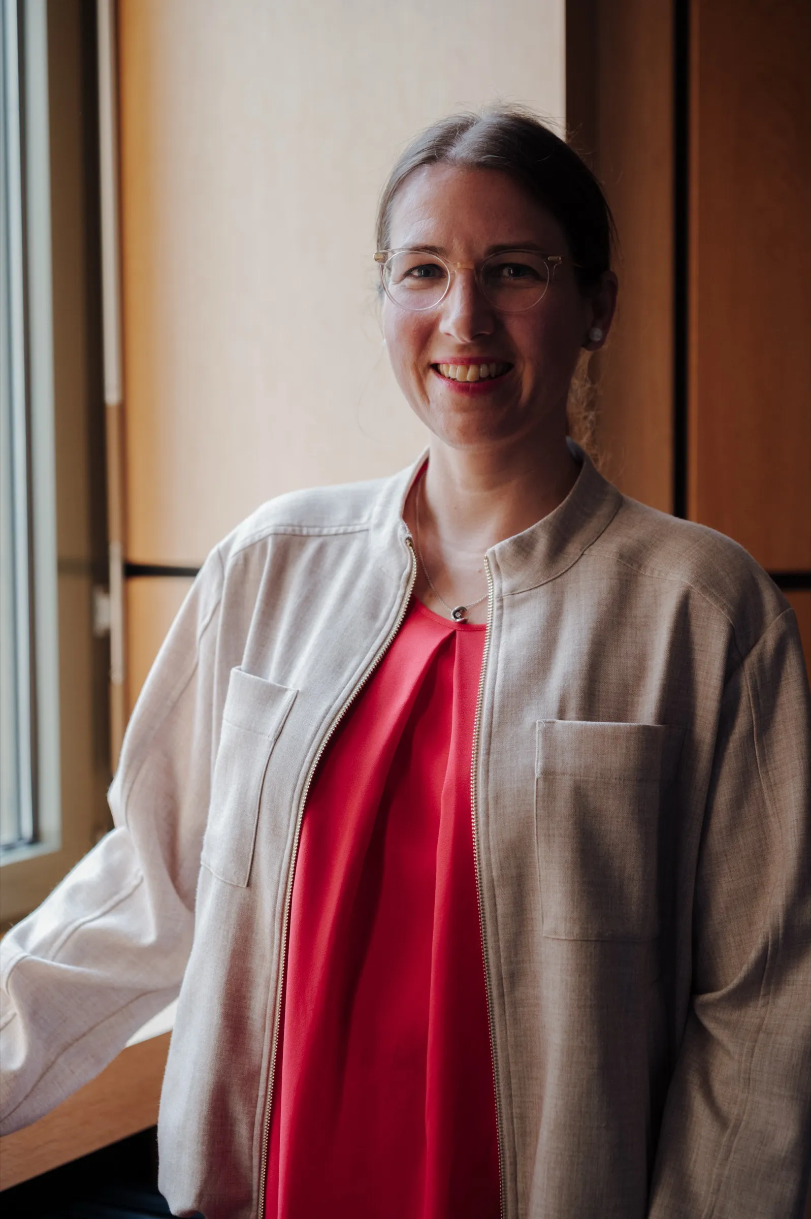 Portrait of Dr. Anna Heinze by the window, red top and light jacket, smiling into the camera