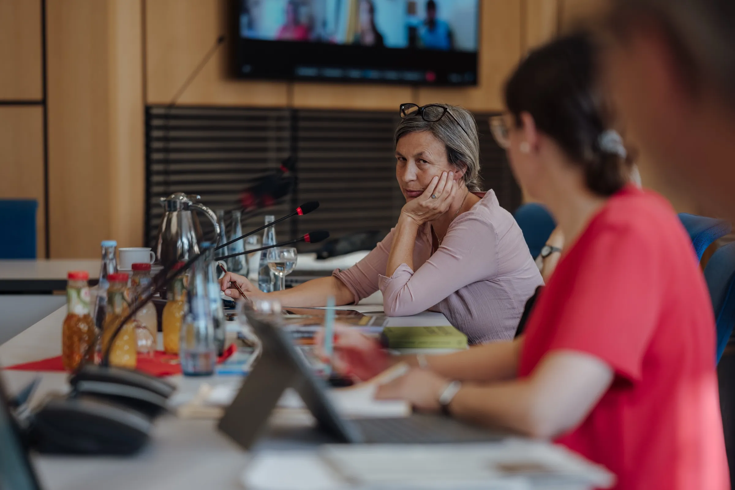Anne Prenzler resting her chin on her hand, listening, Dr. Anna Heinze in red partly in the foreground, on the wall a screen with jury members joining via Teams