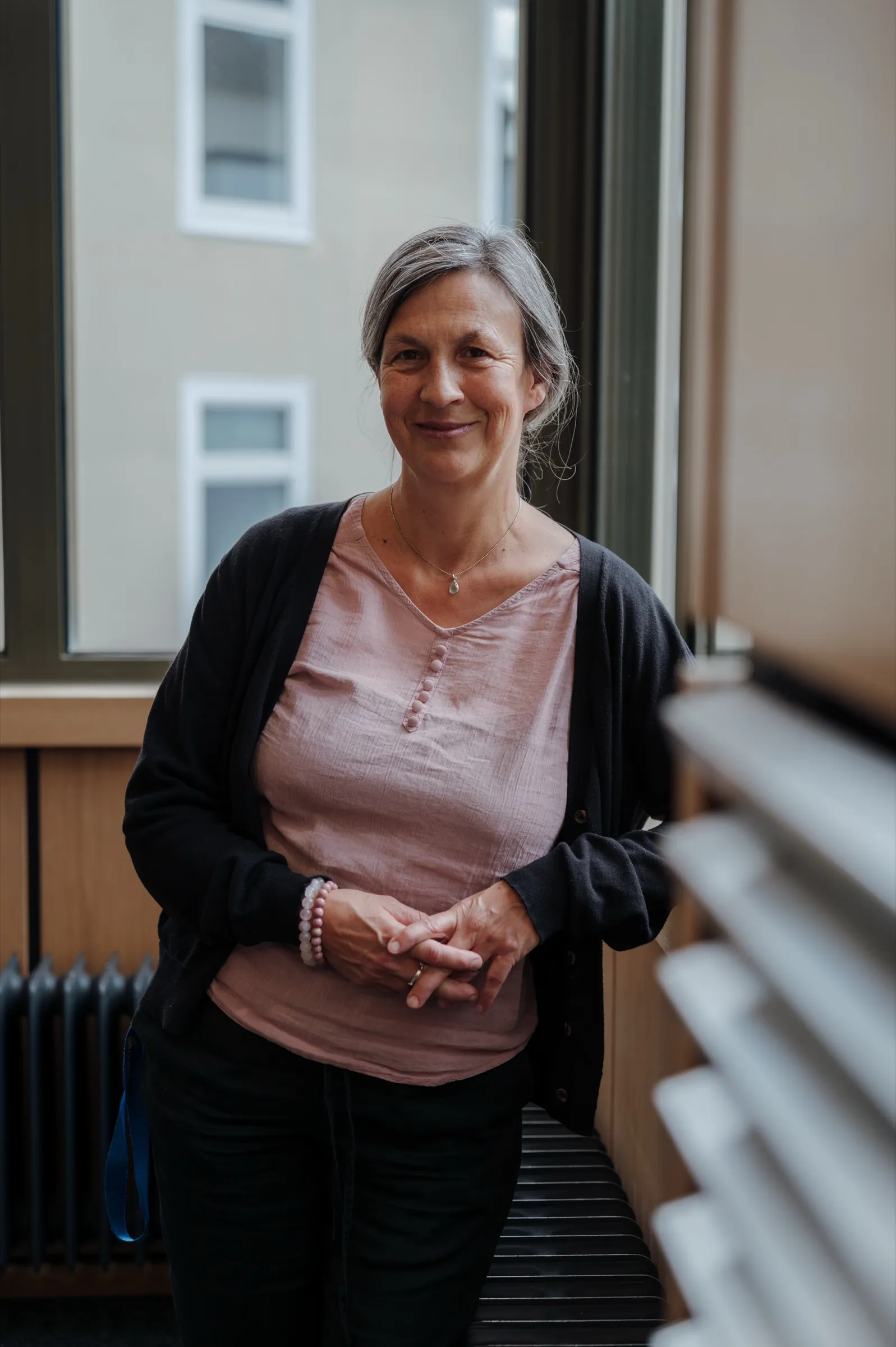Portrait of Anne Prenzler in front of the window, pink blouse and black cardigan, hands loosely folded