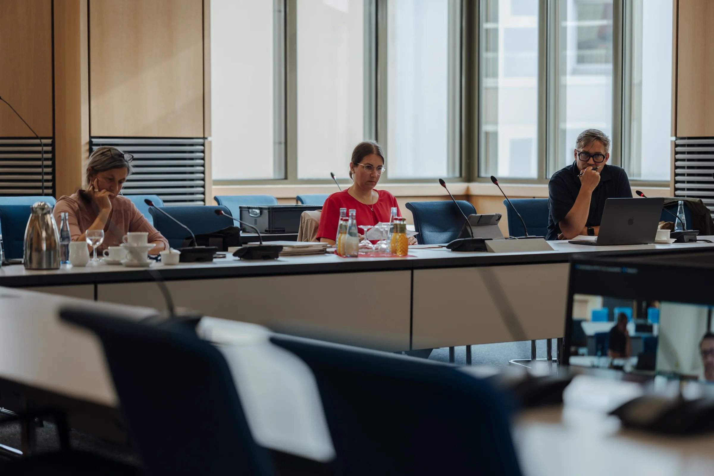 The session room in the state parliament: long white tables with microphones and bottles, blue chairs, tall windows, three jury members at the table