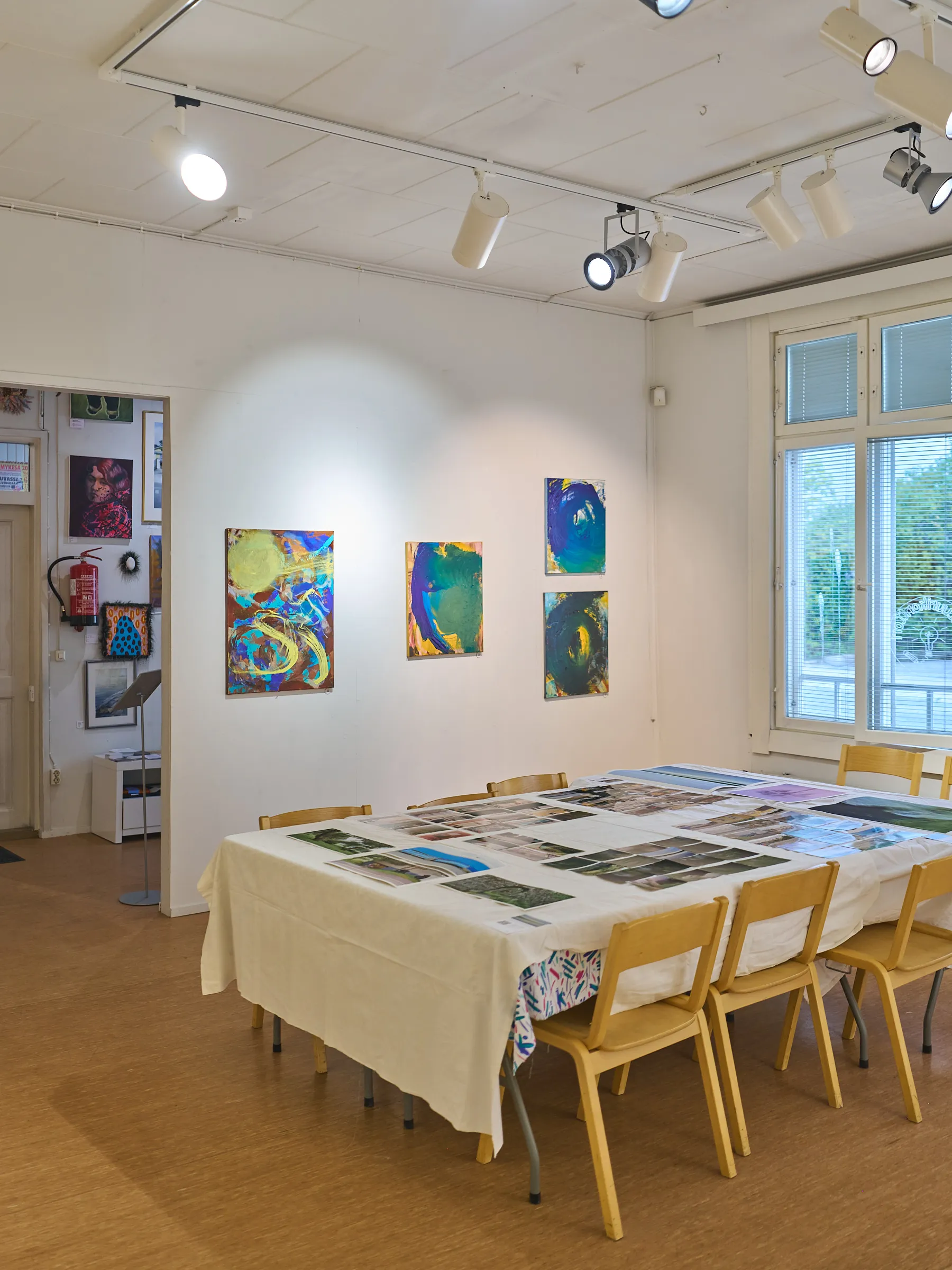 Bright exhibition room with track lights, colourful paintings on the wall and a long table with prints laid out