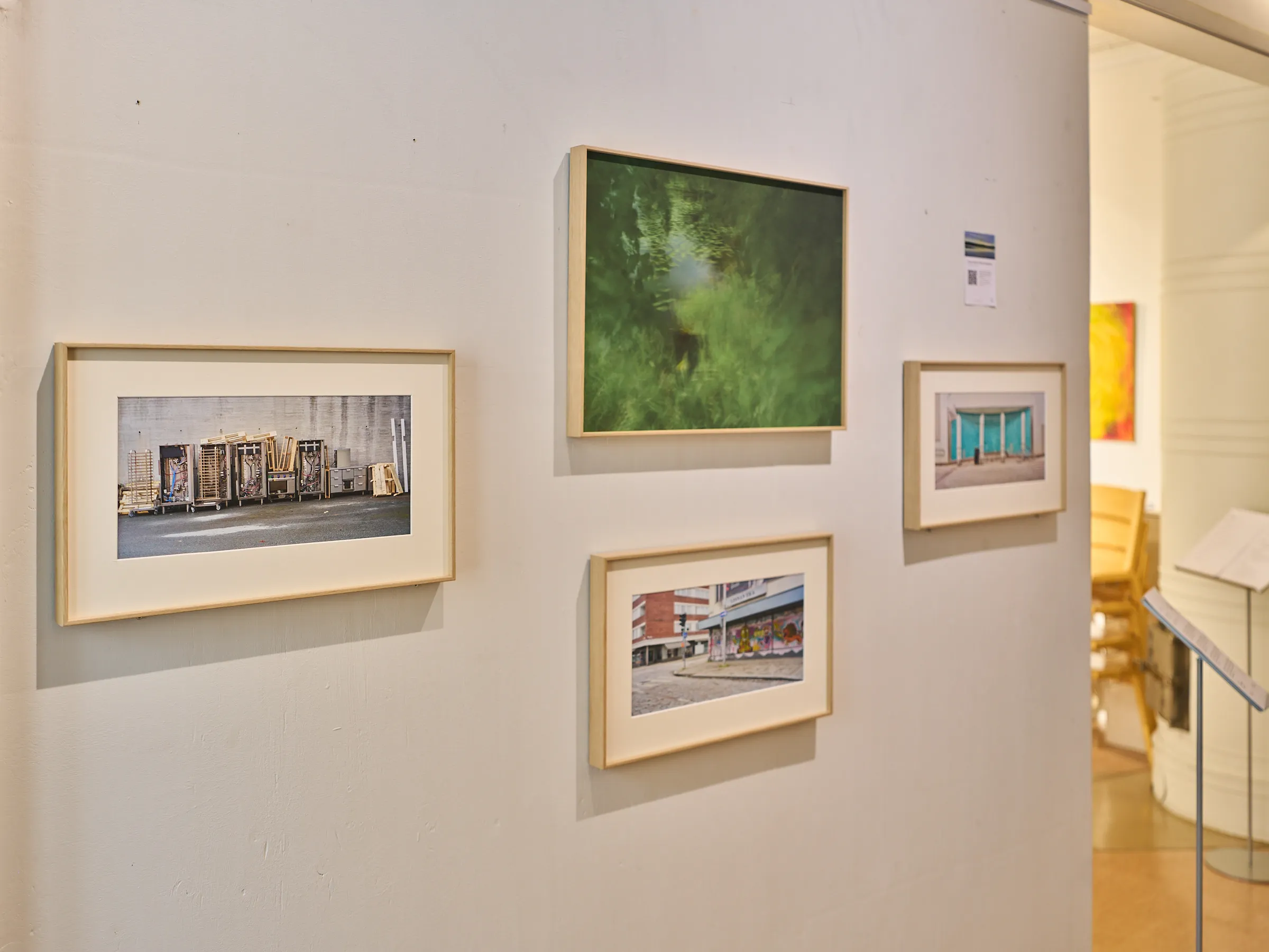 View along the gallery wall, framed photographs and beyond them the bright room with yellow chairs