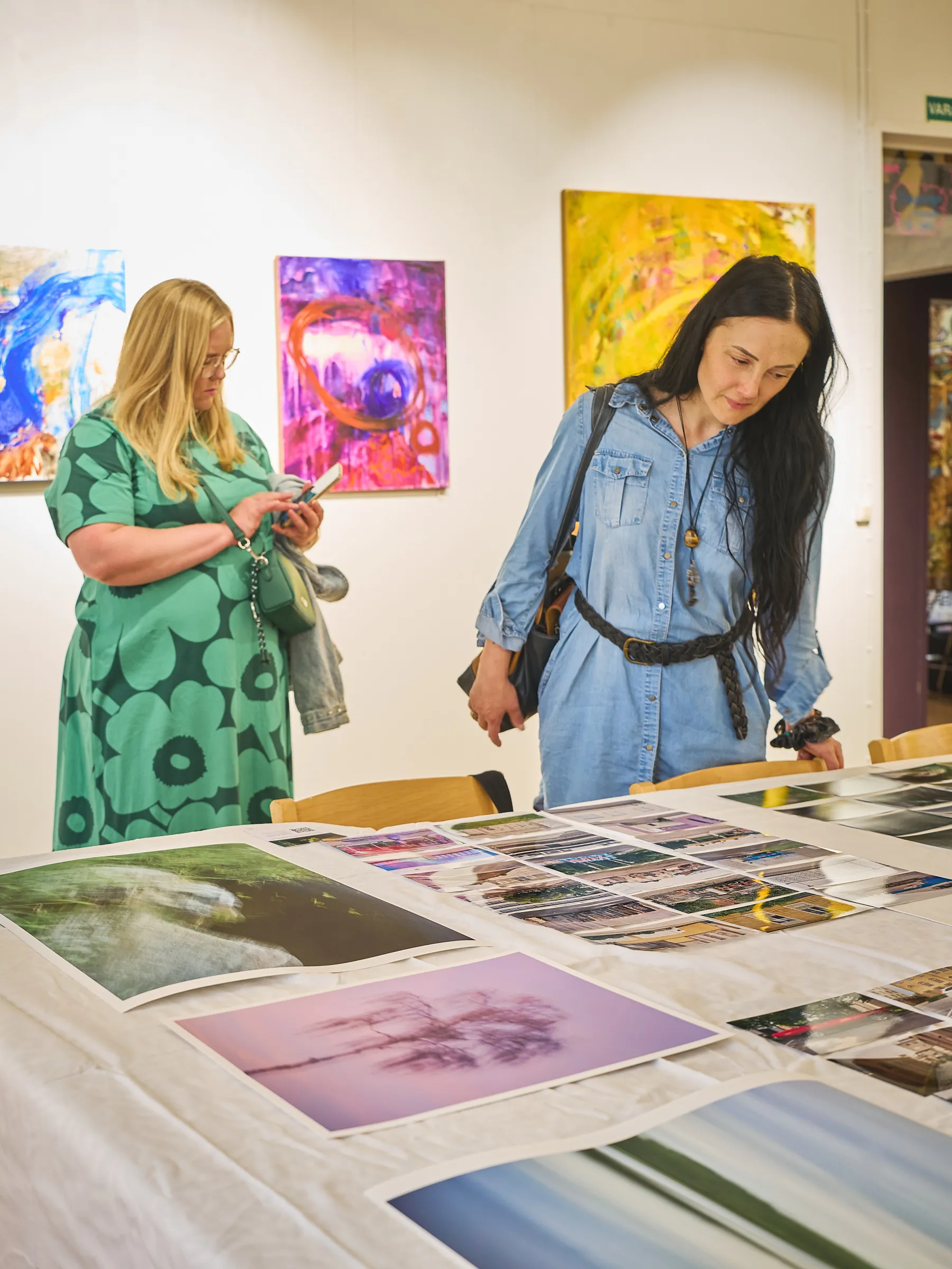 Two women standing in front of colourful paintings, looking at the pictures on the table