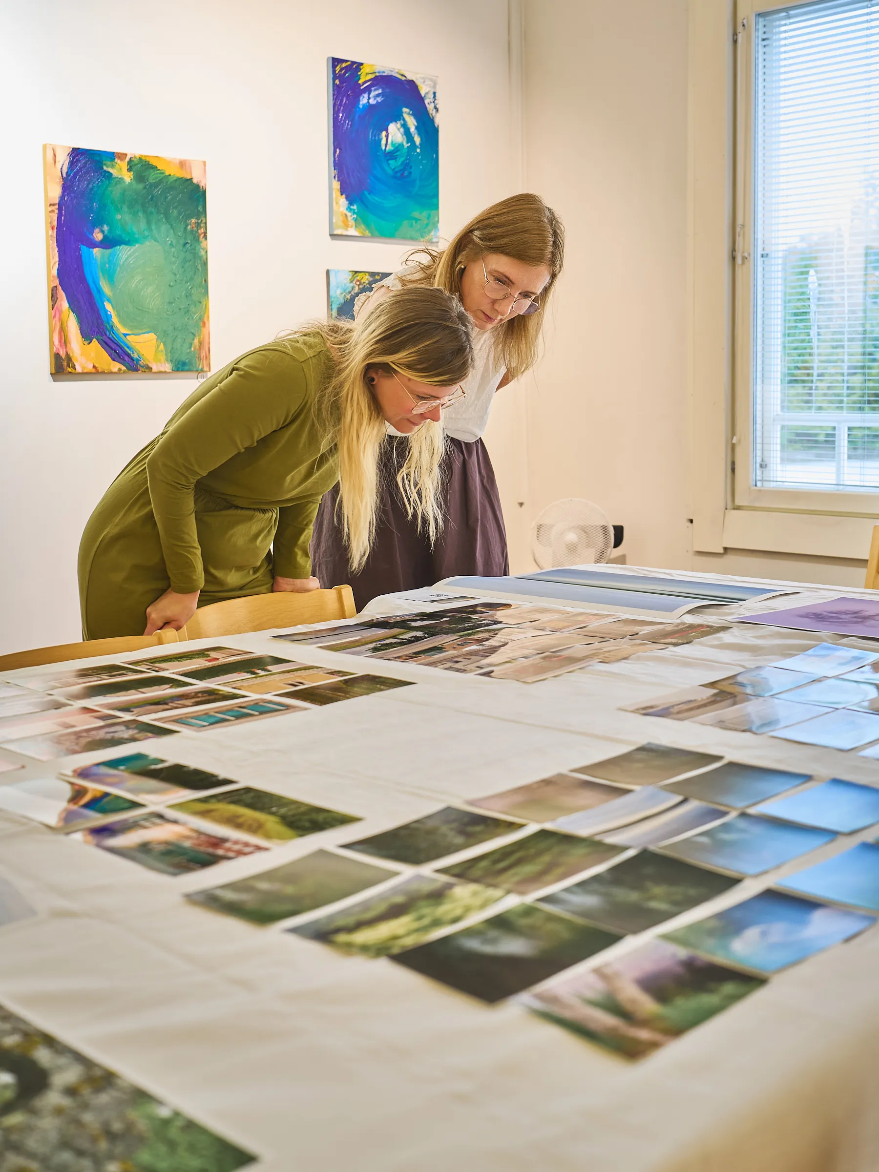 Two women leaning closely over the pictures on the table, colourful paintings in the background
