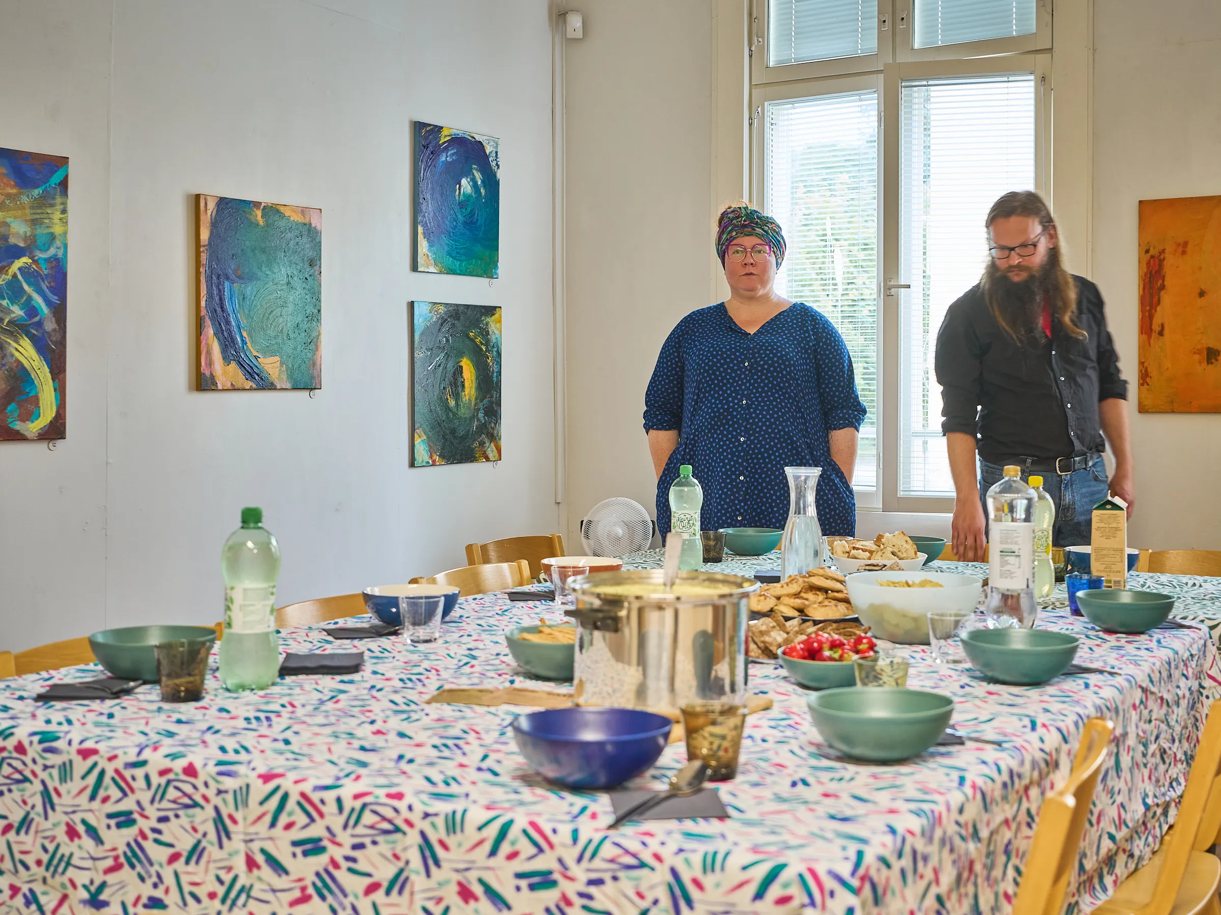 Laid table with a colourful cloth, bowls and bottles, two people standing behind it in the bright room