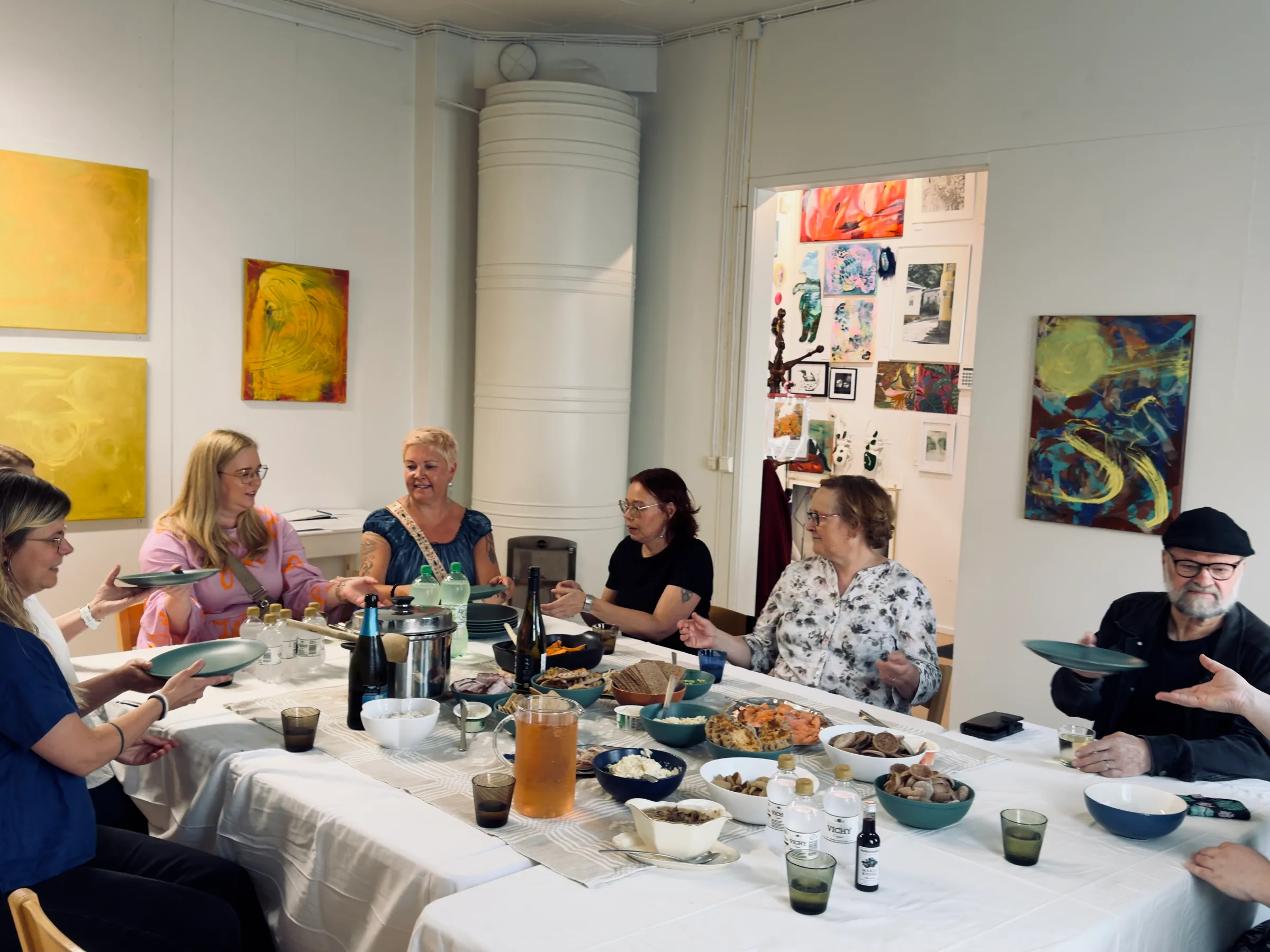 A shared meal at a long table in the Kulttuurikonttori gallery room, paintings on the walls