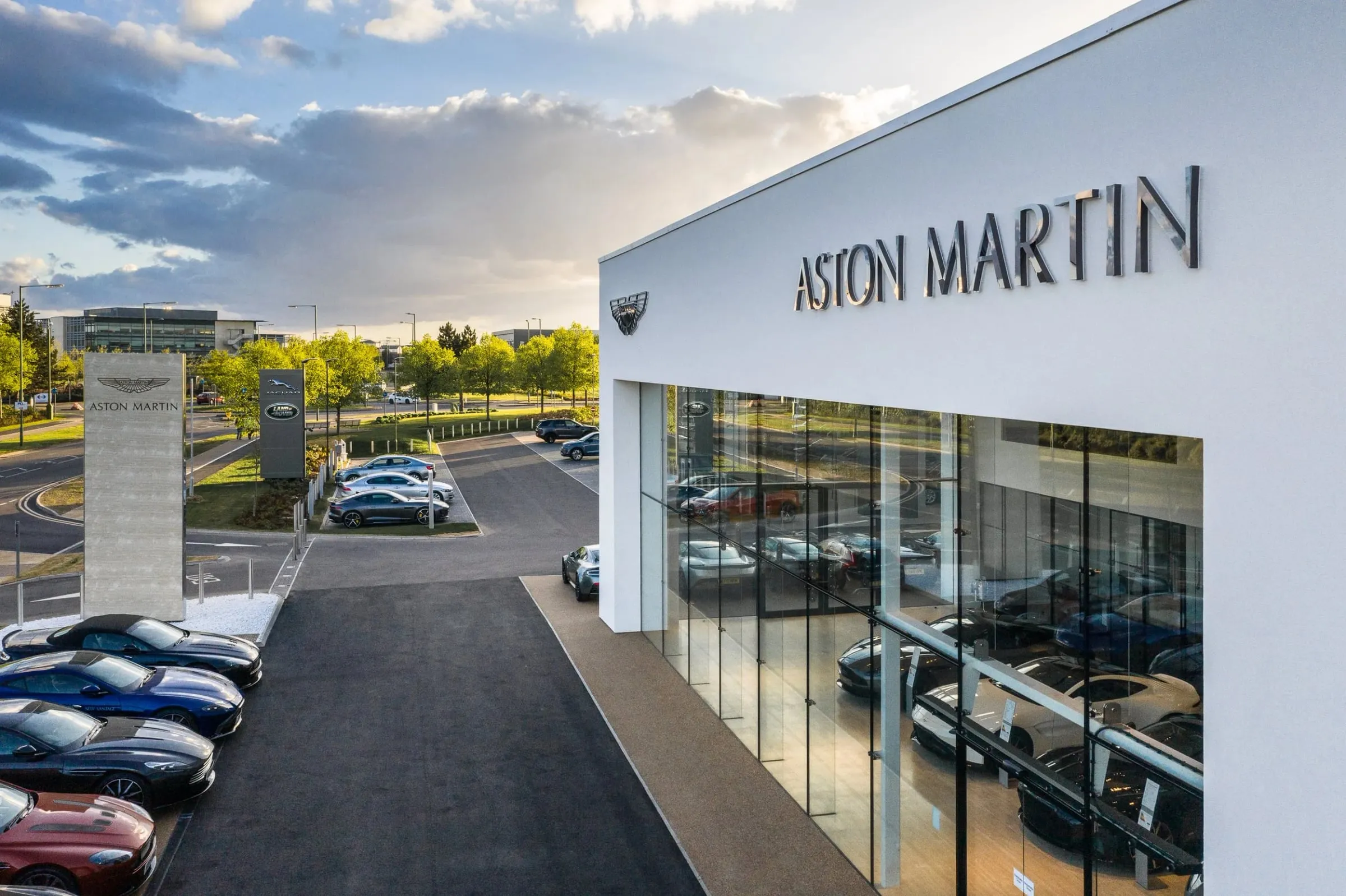Außenaufnahme des Aston Martin Showrooms in der Abenddämmerung, weiße Fassade mit Schriftzug, davor parkende Fahrzeuge