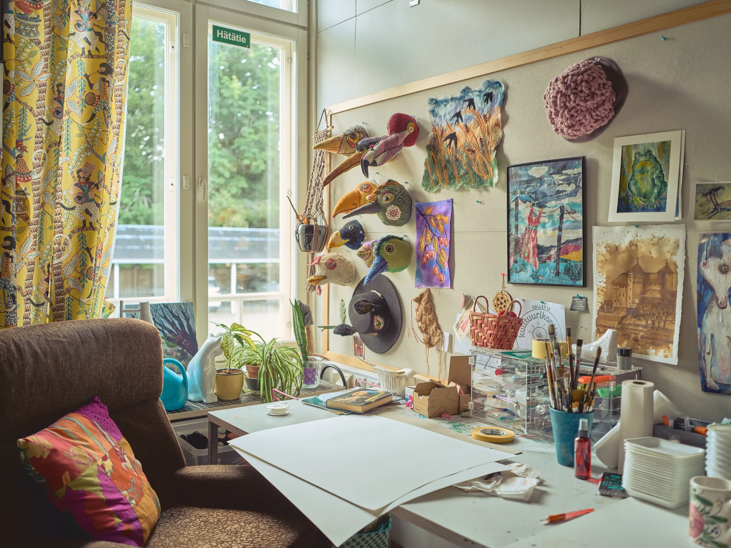 Studio at Kulttuurikonttori: fabric bird masks, felt works and pictures on a pinboard, a work table with brushes in front