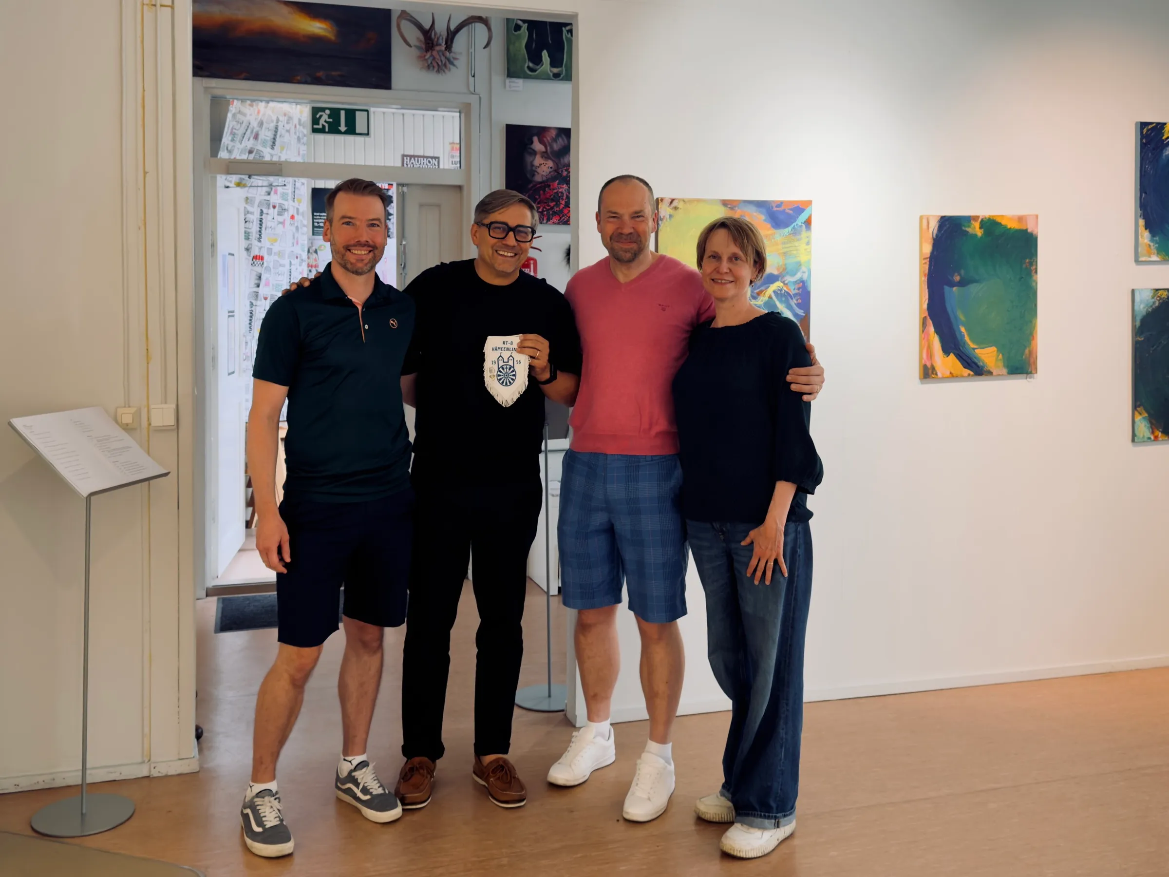 Four people standing arm in arm in the small exhibition, one holding the RT-8 Hämeenlinna pennant